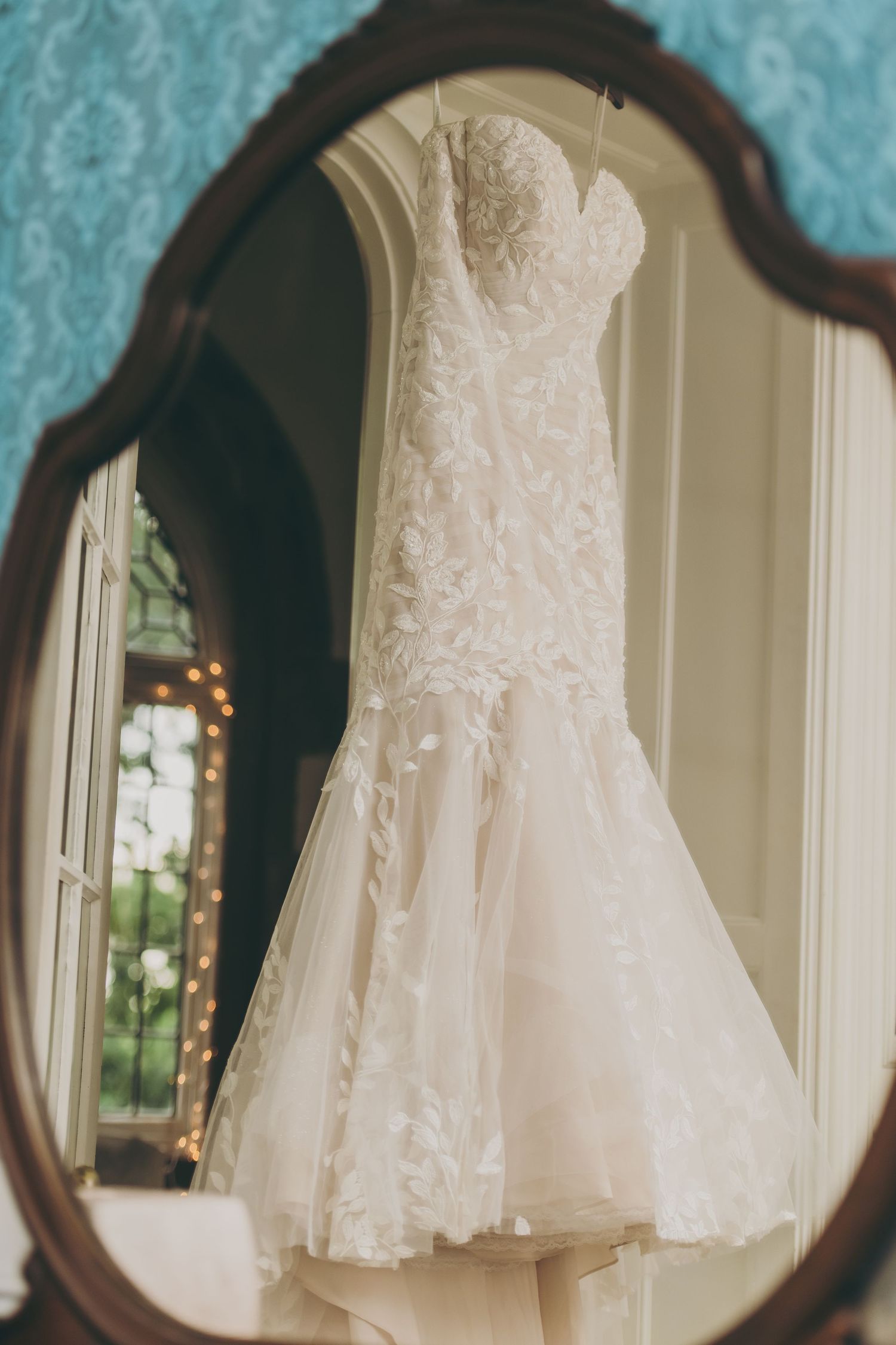 A delicate white lace wedding dress hangs elegantly displayed in front of an ornate mirror.