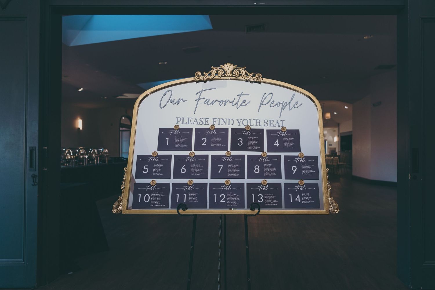 Decorative gold-framed seating chart for wedding guests displayed on dark wall with numbered table assignments.