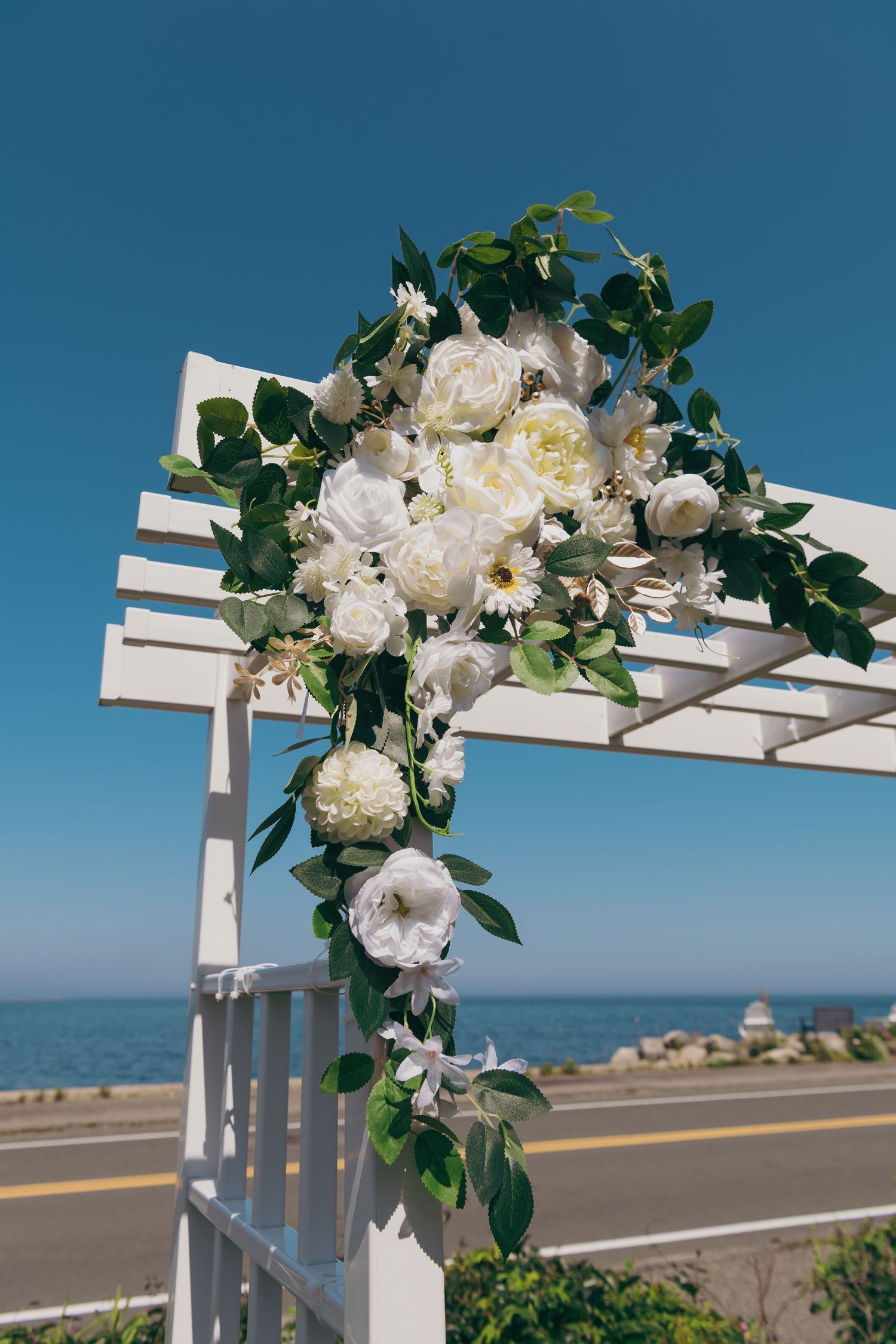White floral arrangement with greenery adorns a coastal pergola overlooking the ocean on a sunny day.