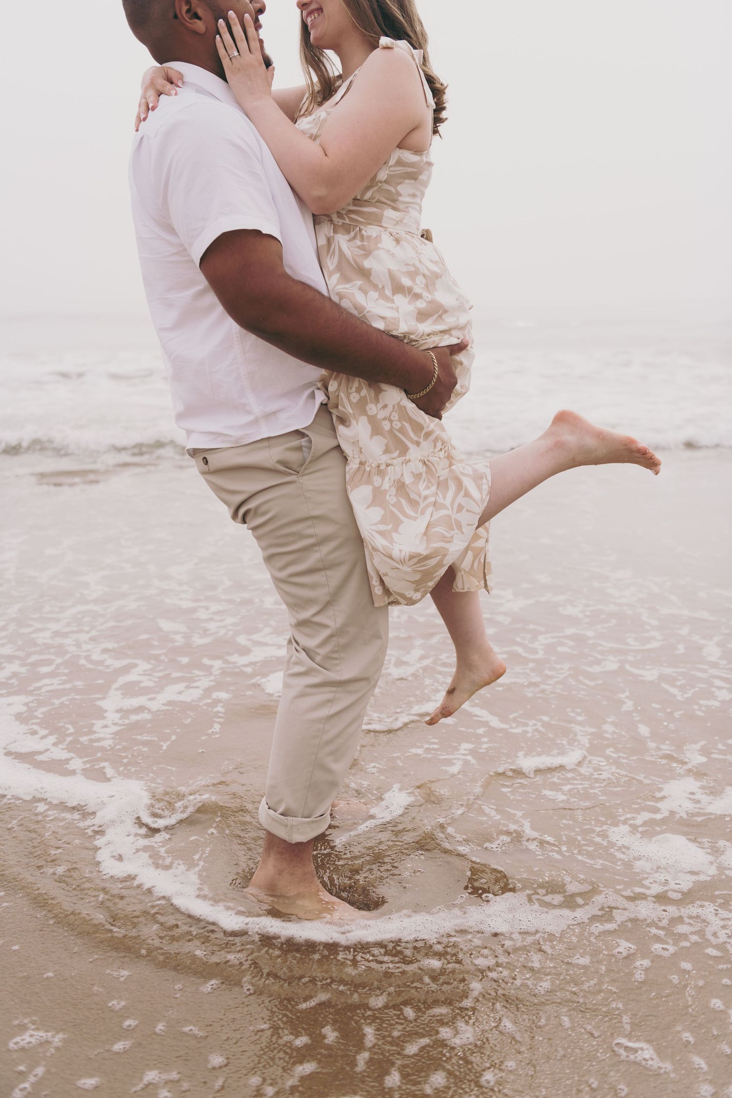 A romantic couple embraces and dances in the shallow ocean waves wearing light summer beachwear on a cloudy day.