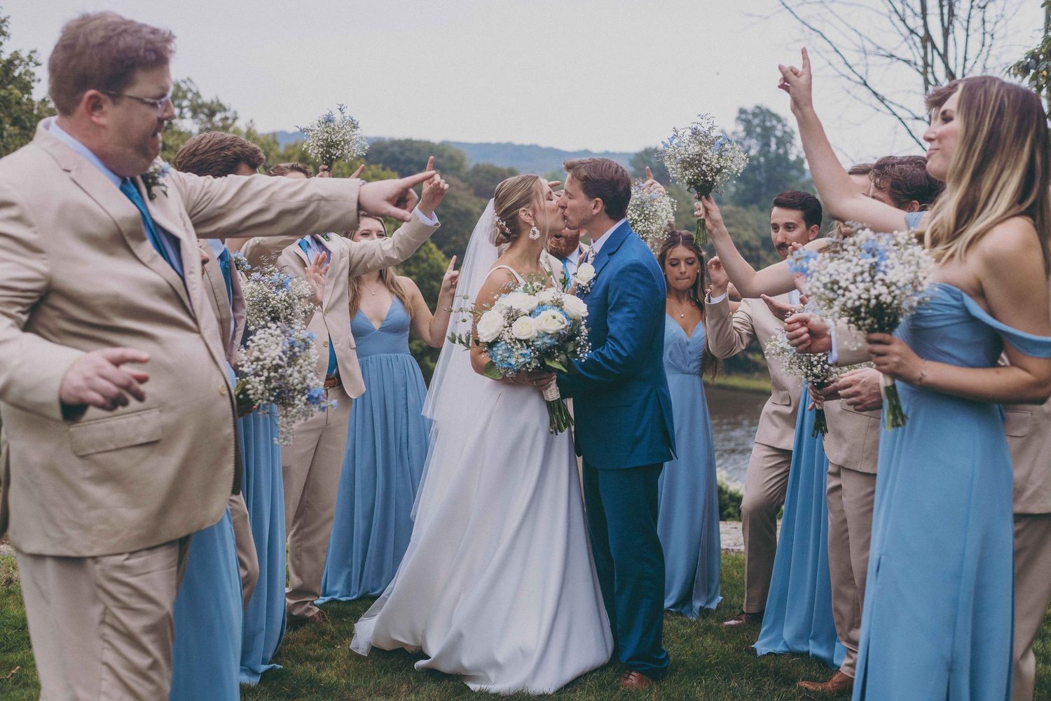 Wedding party in blue dresses and beige suits celebrates outdoors by throwing confetti.