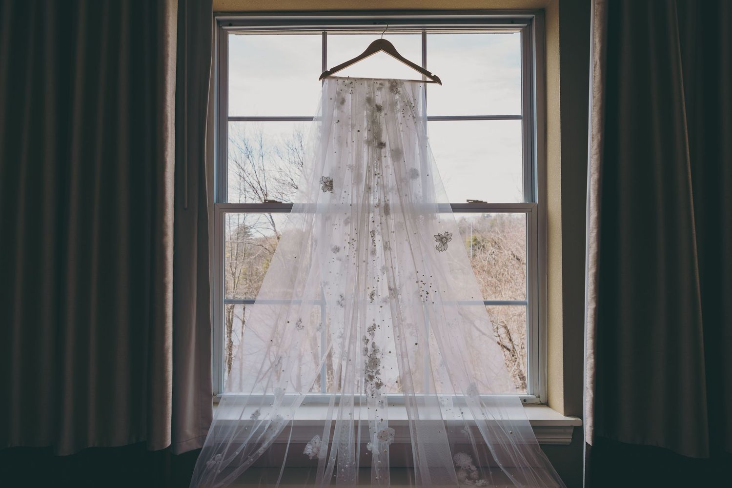 A white lace wedding dress hangs in front of a window with dark curtains creating an ethereal silhouette.