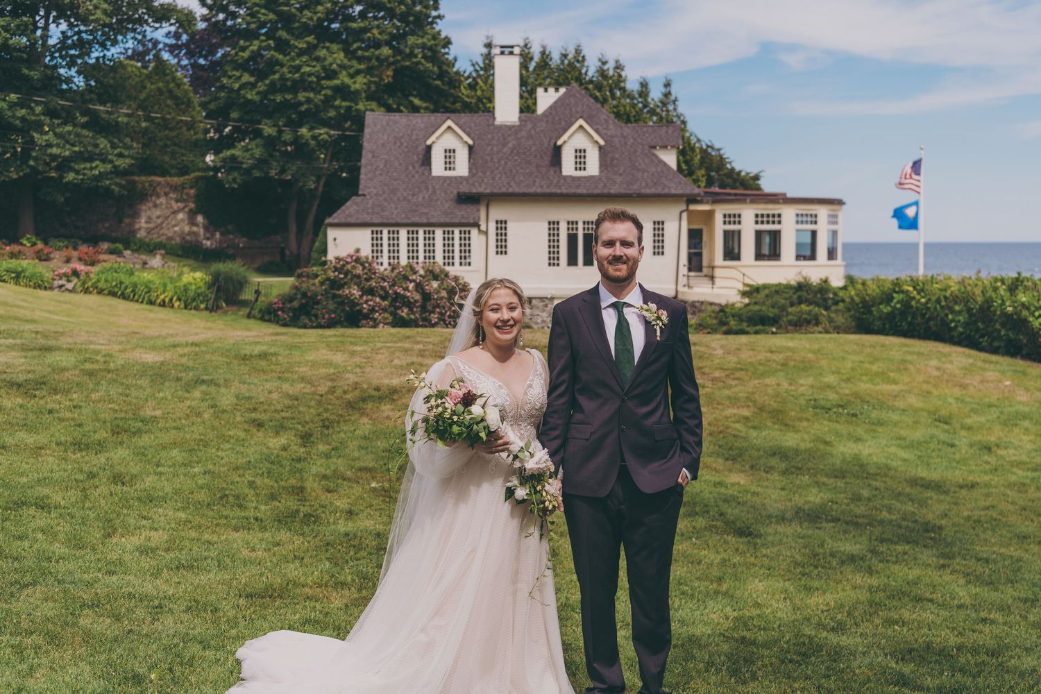 A wedding couple stands on a lawn in front of a beautiful white Cape Cod style house with dormers and Canadian flag.