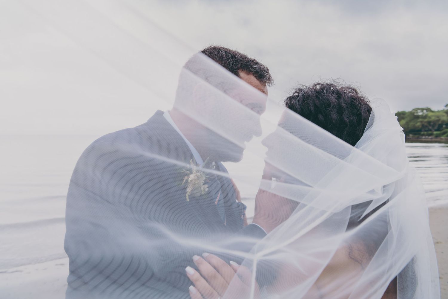 Artistic wedding photo with couple wrapped in flowing veil against cloudy sky.