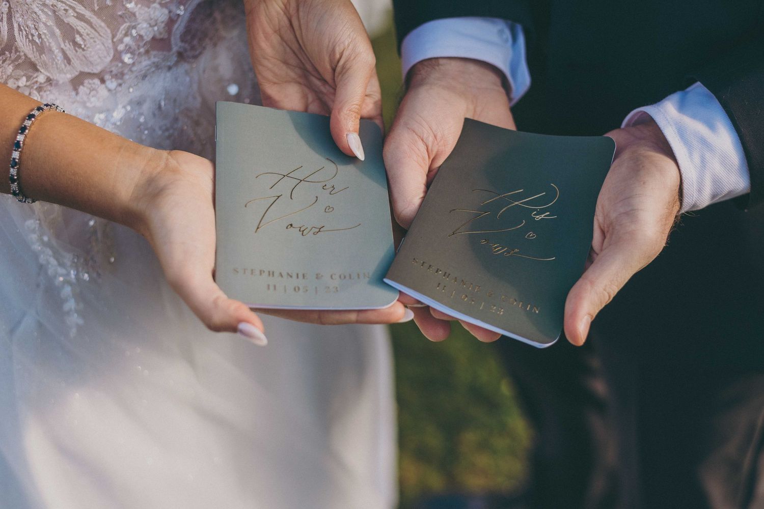 Hands hold matching grey wedding vow books with decorative text during an outdoor ceremony.