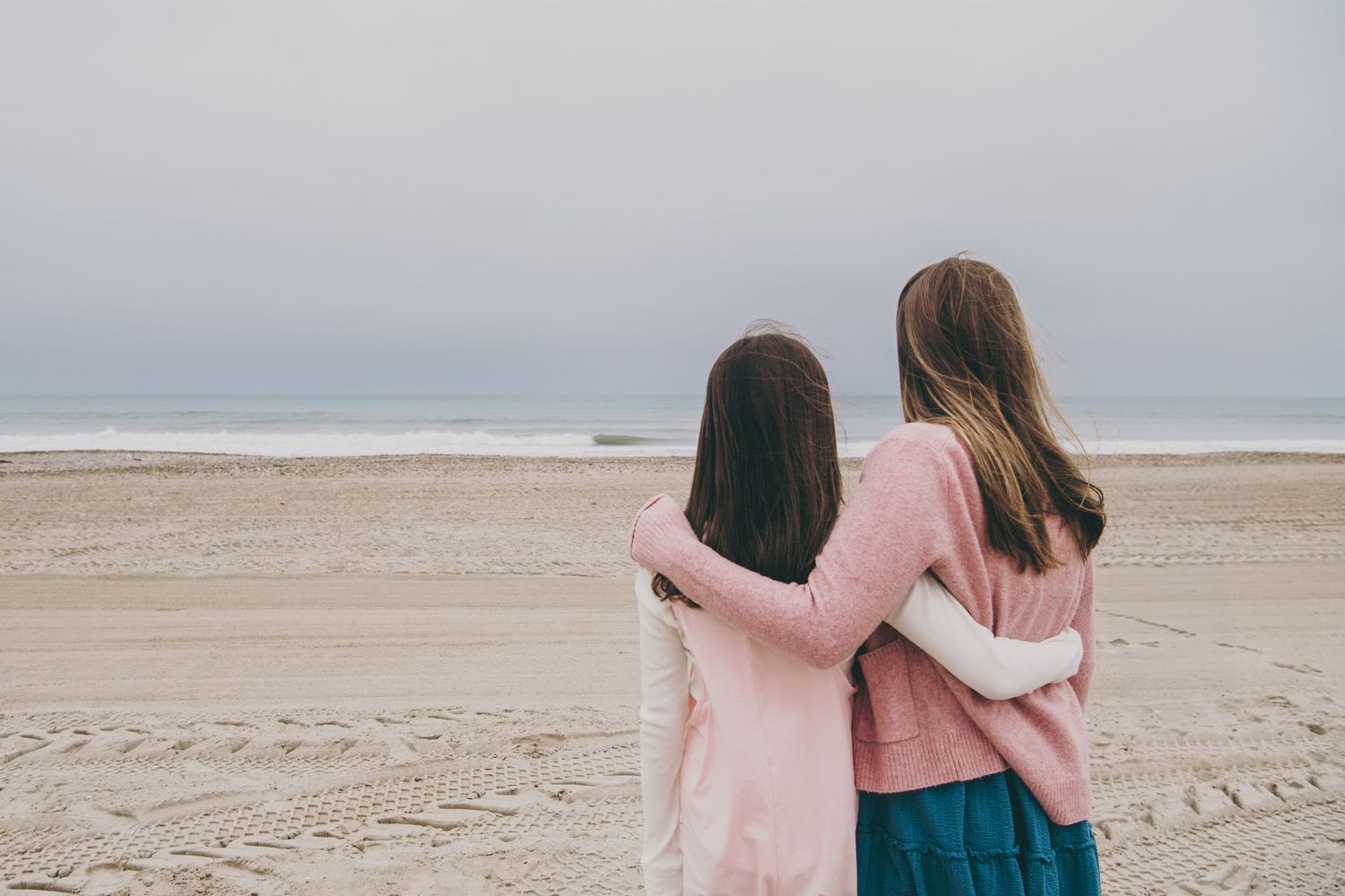 Two friends wearing light pink sweaters embrace while standing on a misty beach.