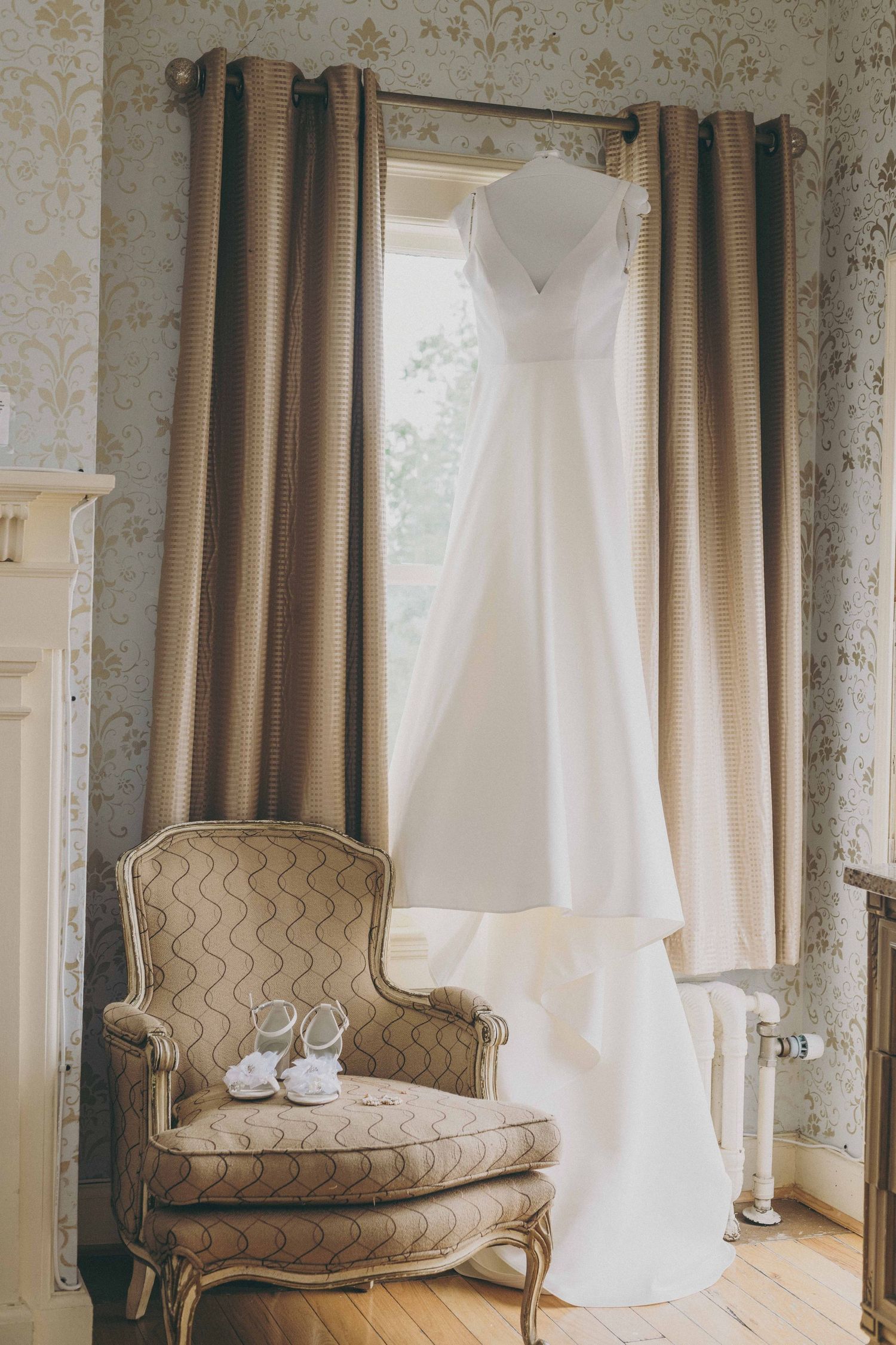 An elegant white wedding dress hangs by a window next to a vintage beige chair with bridal shoes.