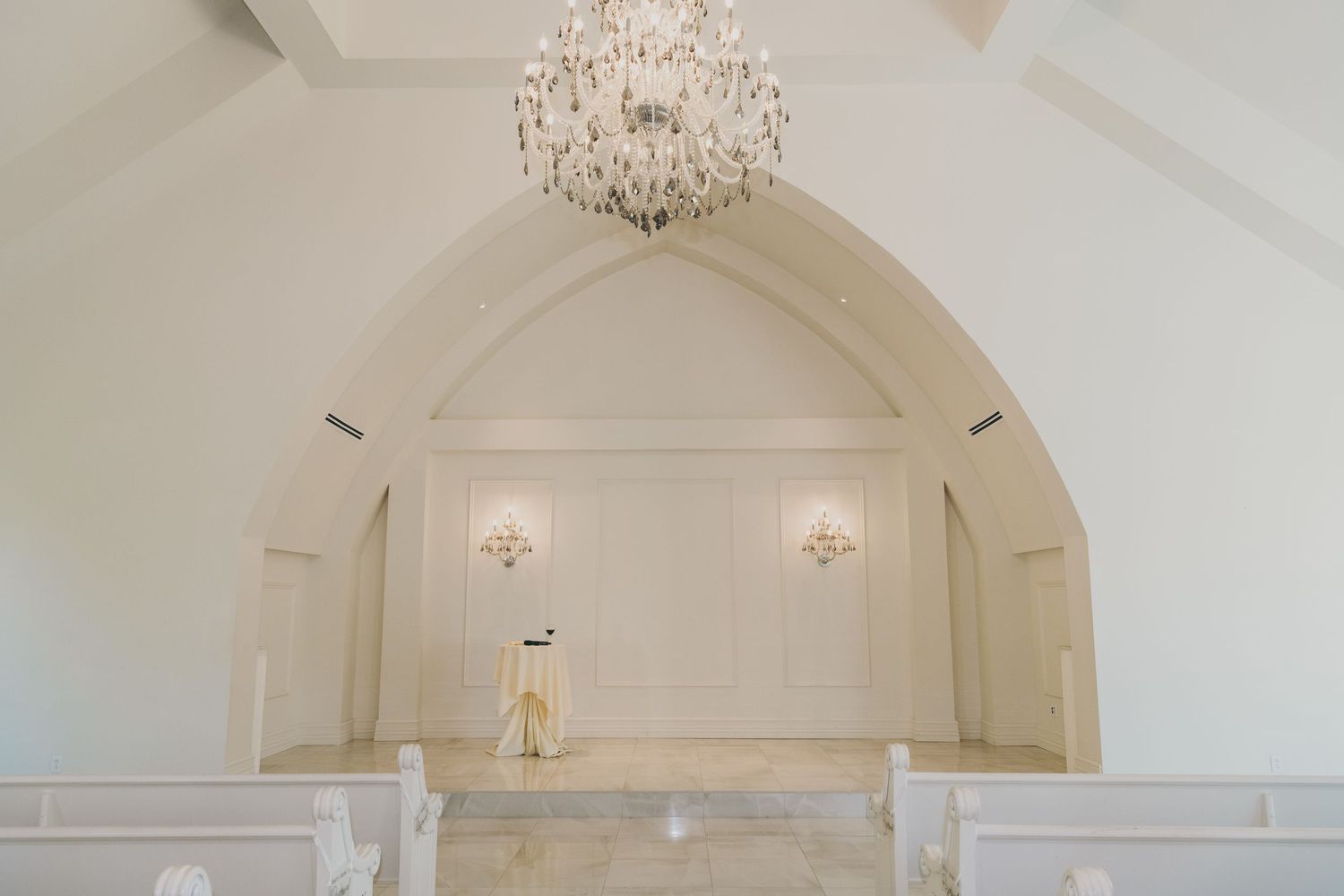 An elegant white chapel interior with arched ceiling, crystal chandelier and wall sconces illuminating the pristine space.