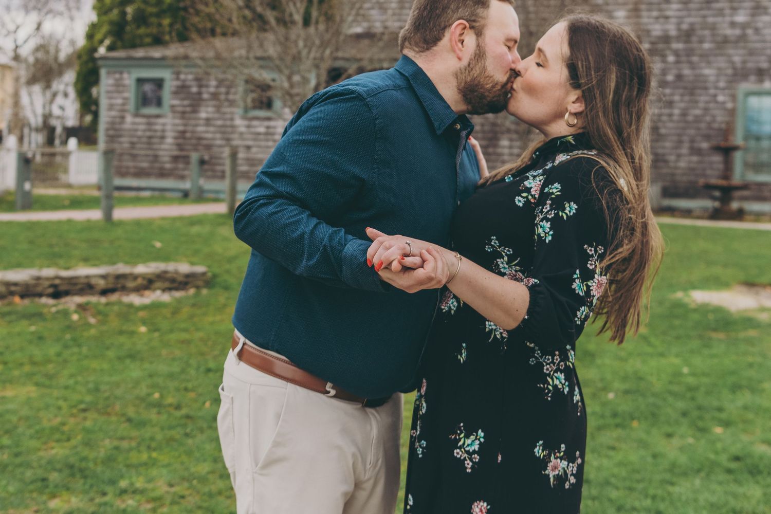 A couple shares an intimate embrace while standing on grass with rustic buildings in the background.
