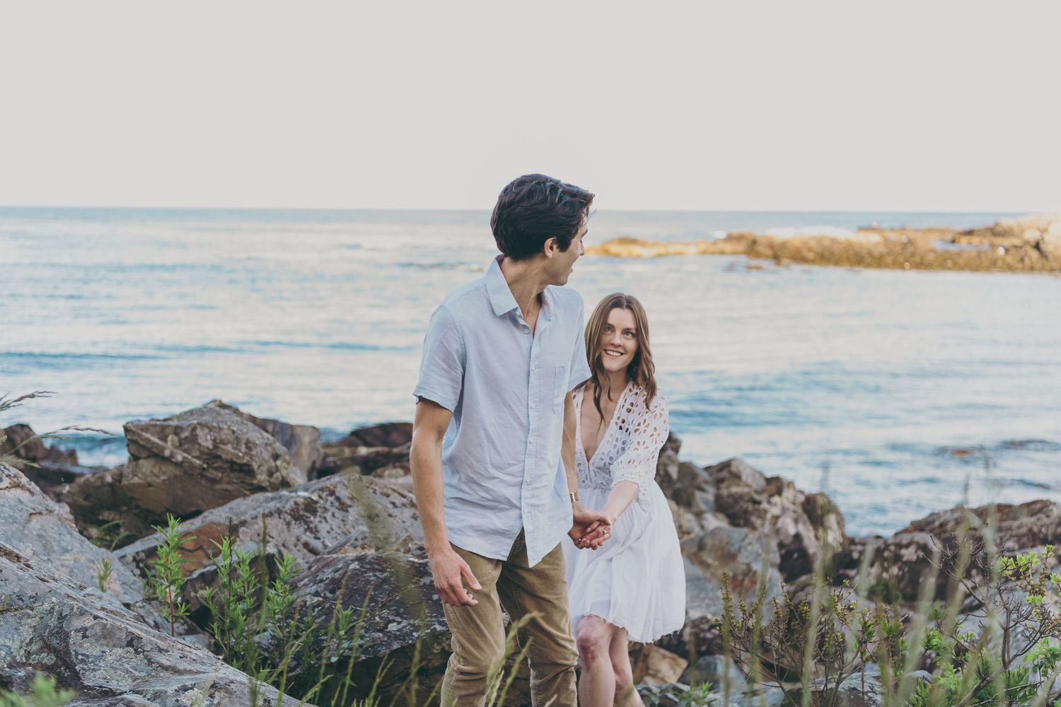 Two people stand together on rocky coastal cliffs with ocean waves and shoreline in background during sunset.