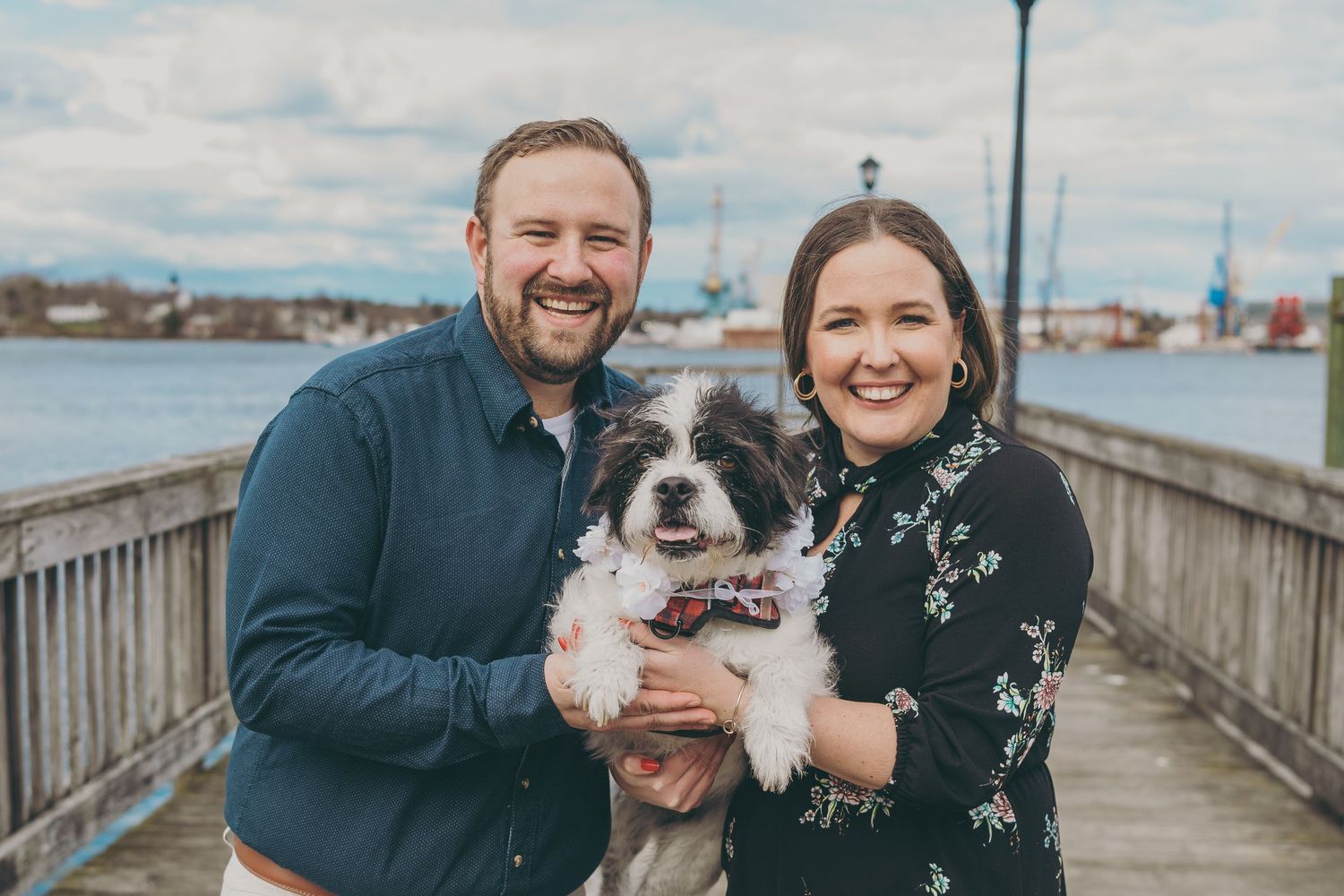 A happy couple poses with their scruffy black and white dog on a waterfront boardwalk.