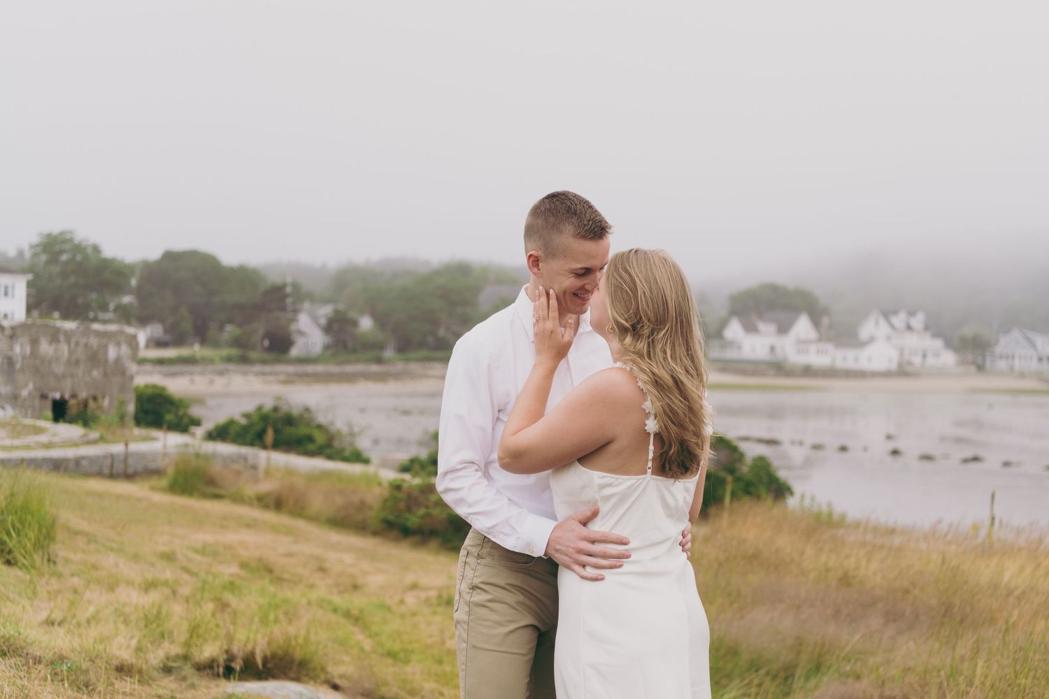 A couple embraces on a misty coastal hillside overlooking a scenic harbor with white houses in the background.
