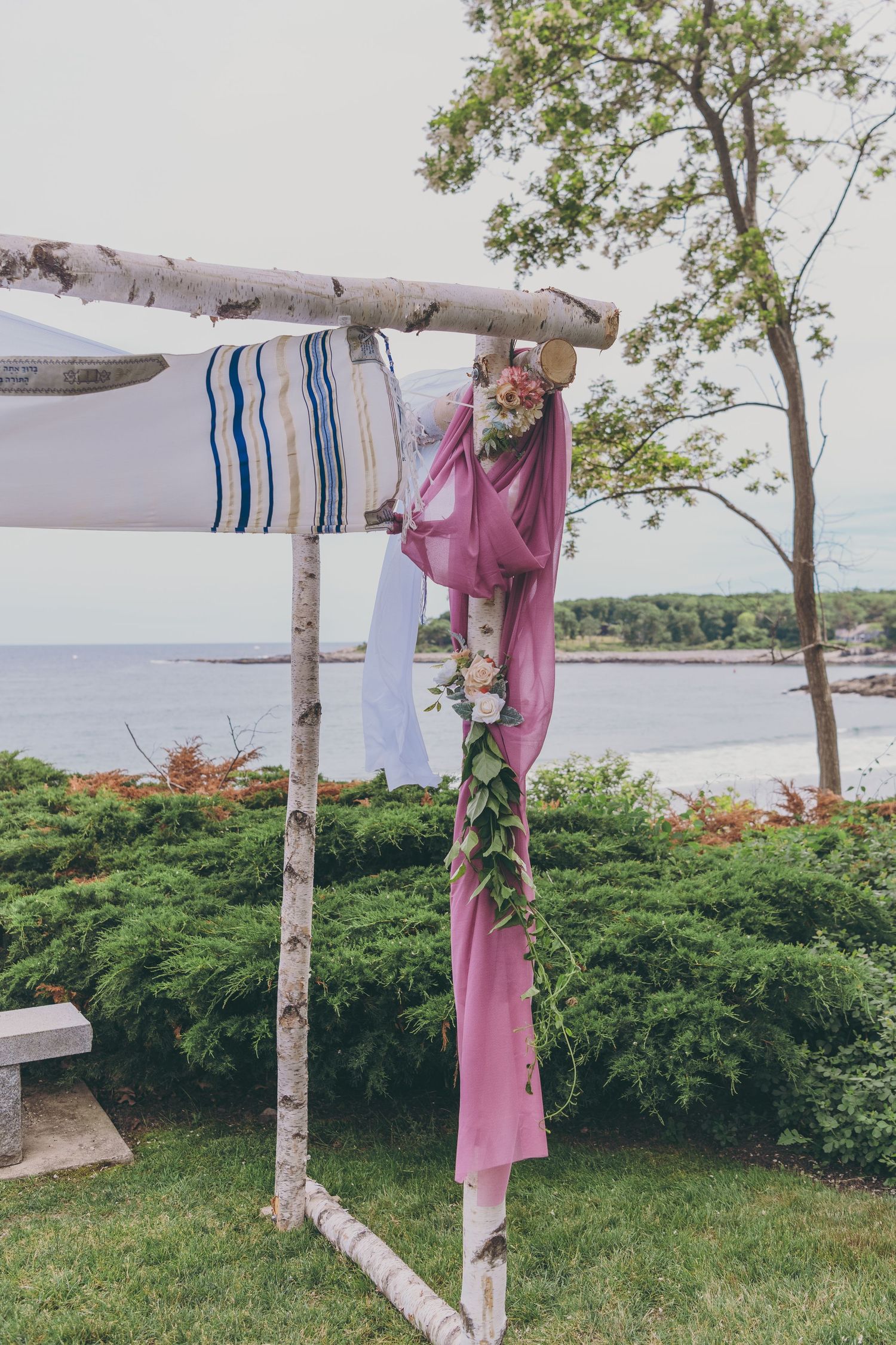 A rustic birch wood wedding arch decorated with pink fabric and floral arrangements overlooks a scenic waterfront.