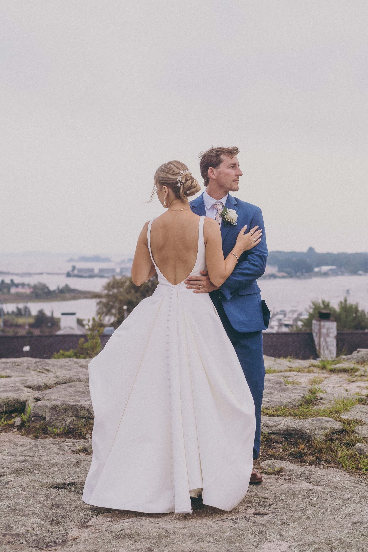 A romantic wedding couple embraces on a clifftop overlooking a misty waterfront landscape.