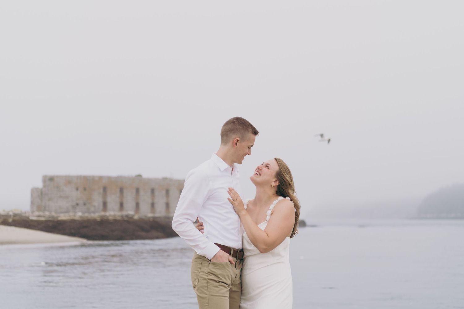 A couple in wedding attire shares a romantic moment on a foggy beach with a stone fortress in the background.