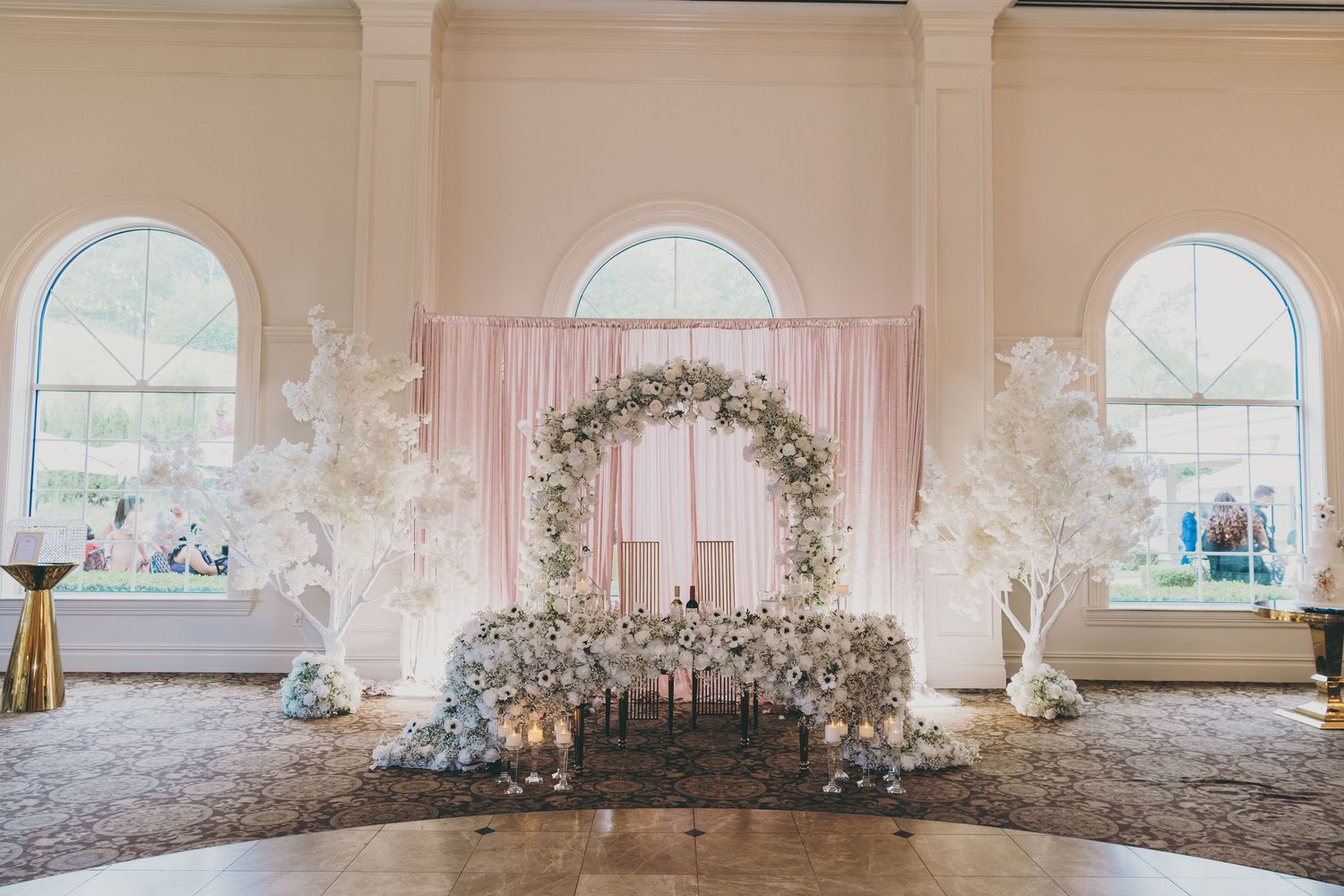 White floral wedding arch decorations in a light pink ballroom with elegant drapery and arched windows.
