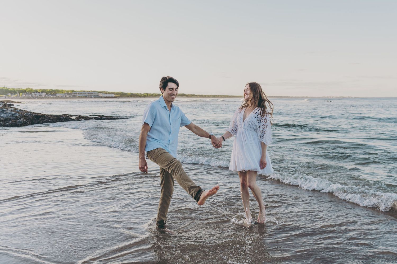 A couple splashes and plays in the shallow ocean water at sunset while wearing casual beach attire.