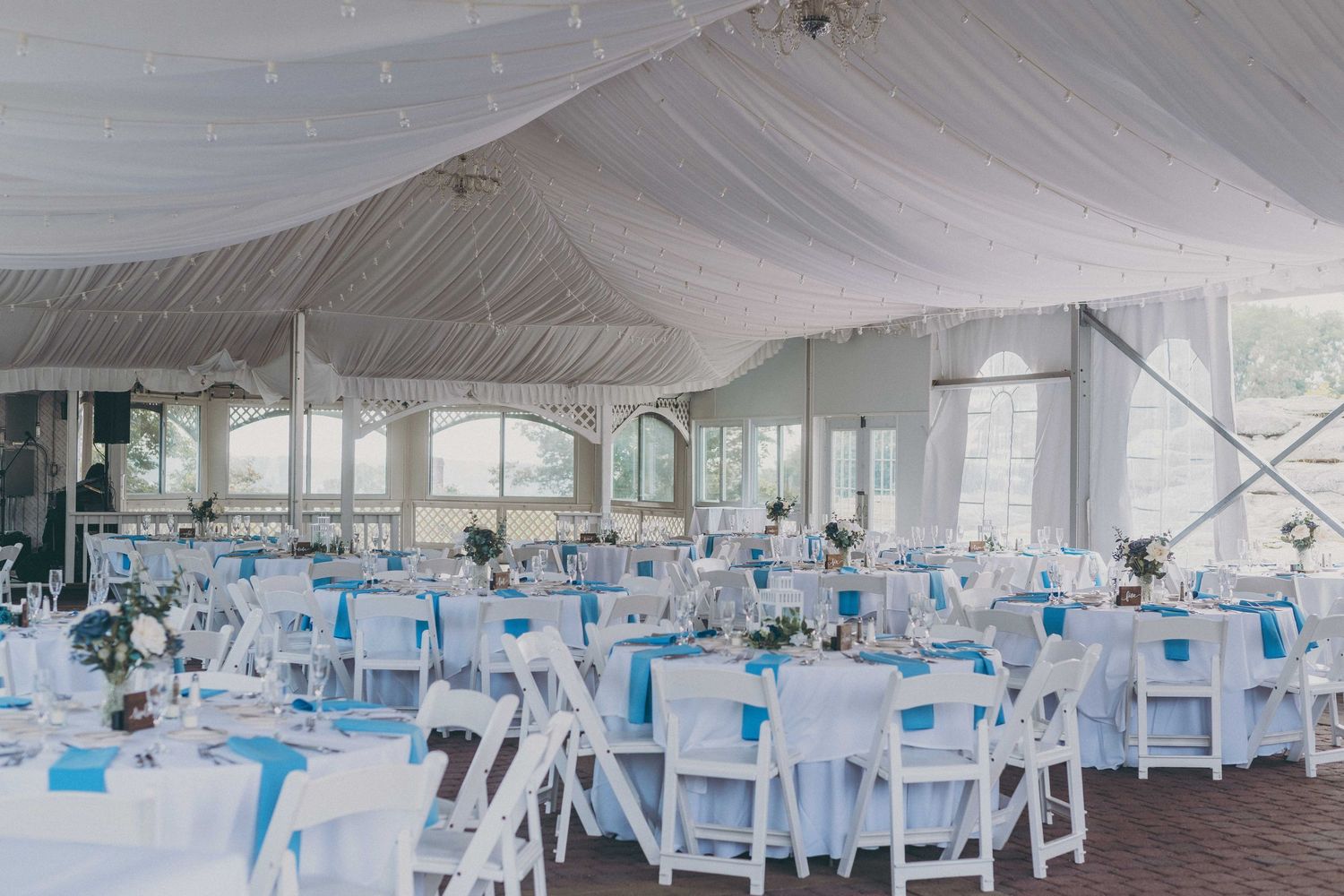 An elegant wedding reception venue with white draped ceiling, round tables, and turquoise accents under a large tent structure.