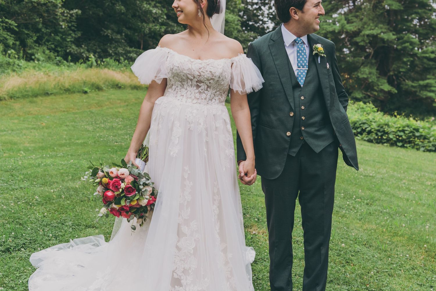 A couple stands together at their outdoor wedding, with the bride in a lacy white dress holding a vibrant red bouquet.