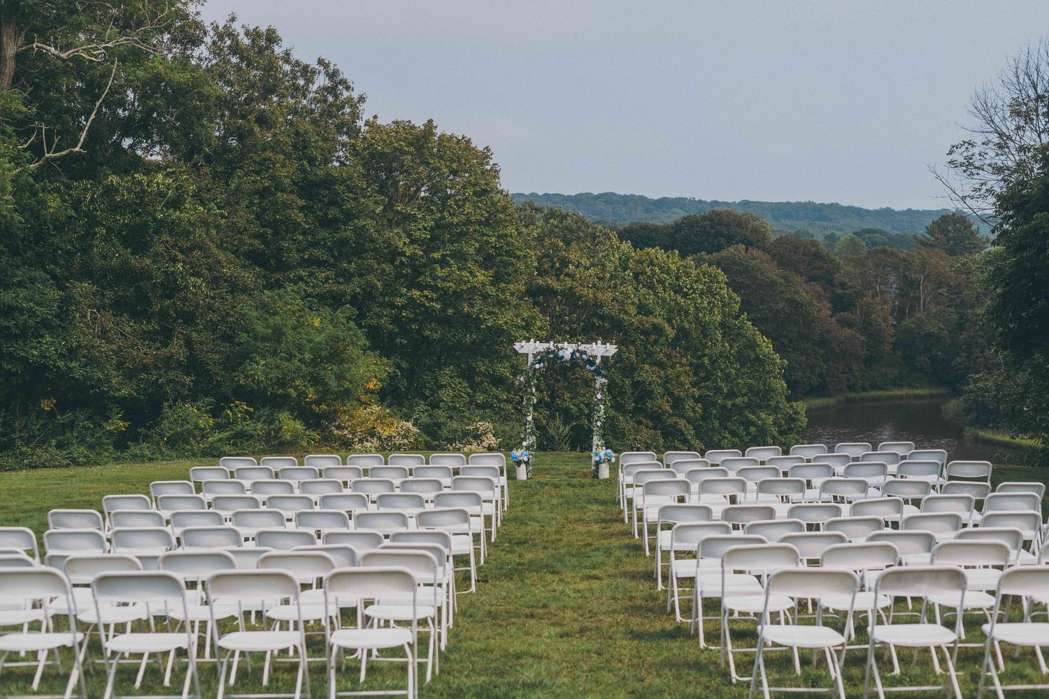 White folding chairs arranged on a grassy hillside for an outdoor wedding ceremony with mountains in the background.
