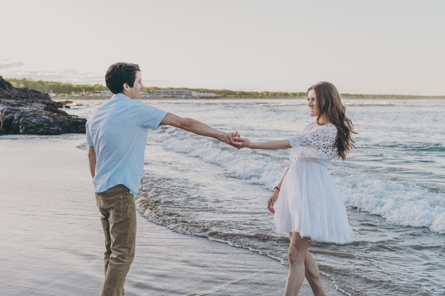 A romantic couple holding hands and walking along a sandy beach at sunset with waves gently rolling in.