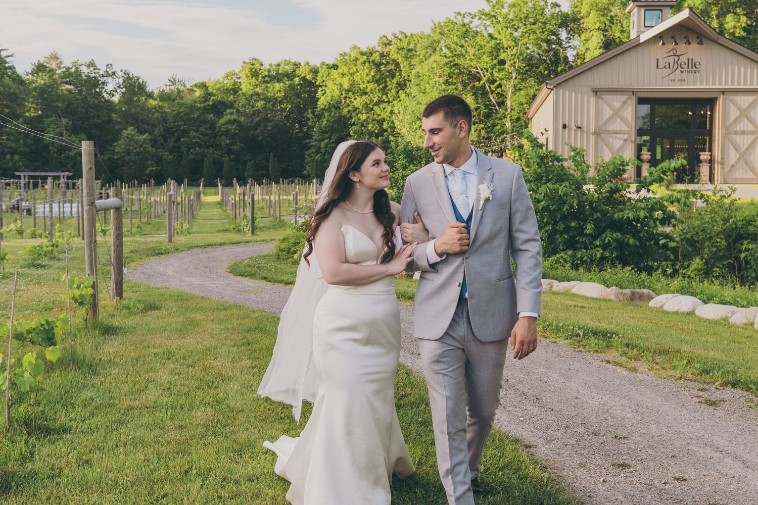 Couple embracing outdoors at rustic vineyard venue with wooden barn and rolling hills in background.