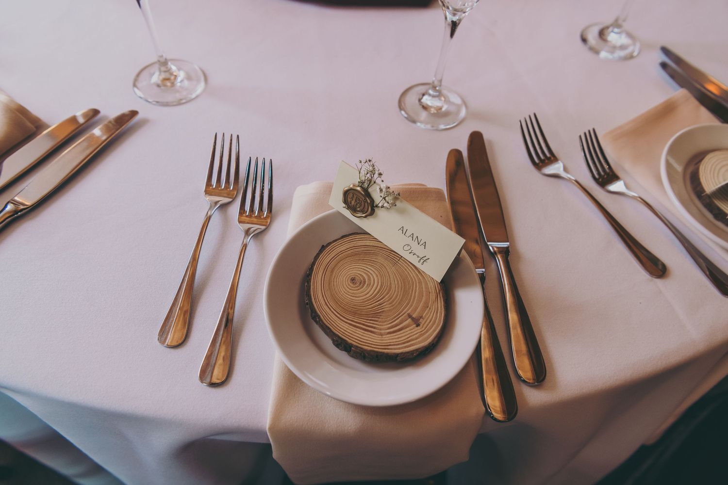 Place setting at wedding reception with wooden coaster, silver utensils, and wine glasses on white tablecloth.