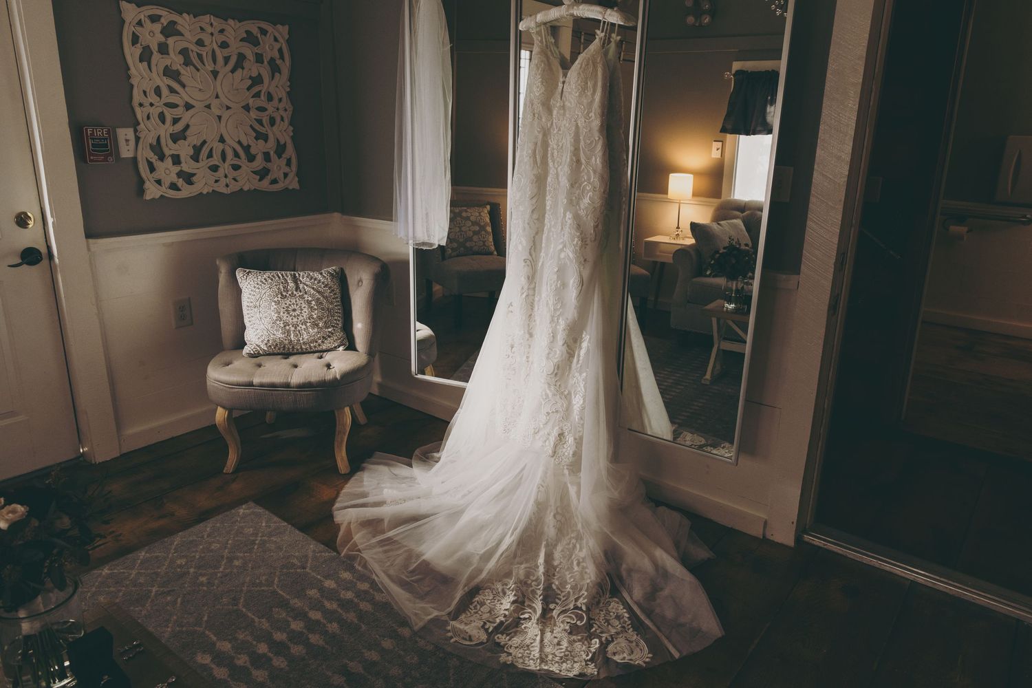 An elegant white lace wedding dress hangs in a dimly lit room with a vintage chair and decorative wall art.