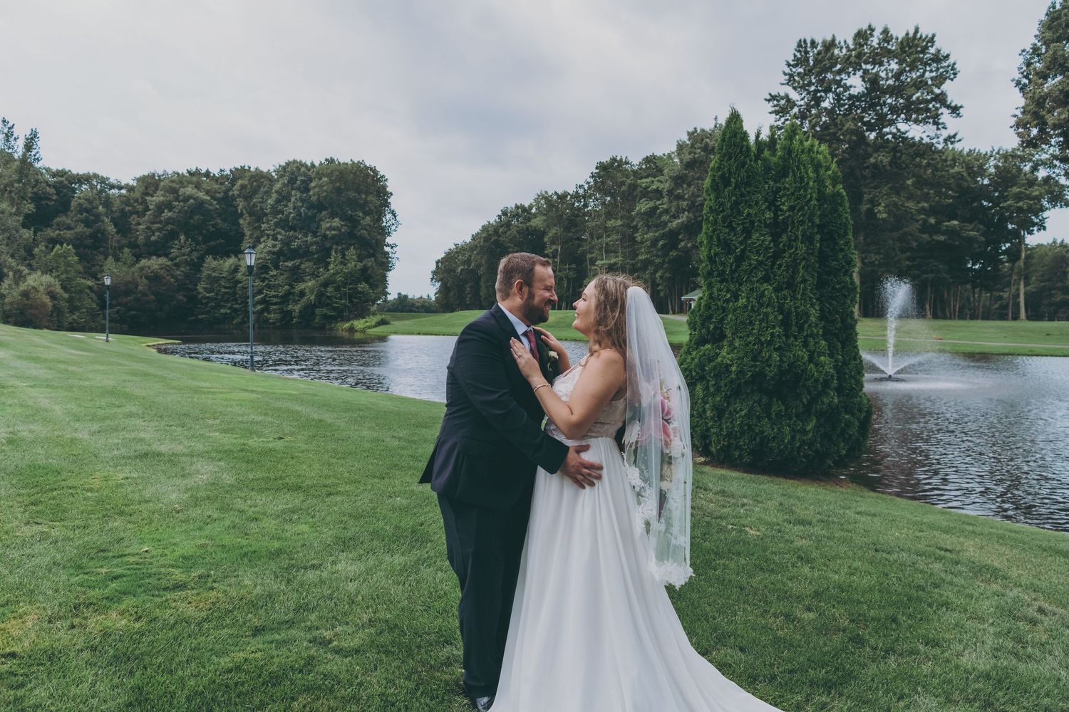 A romantic wedding moment captured on a manicured lawn beside a peaceful pond with evergreen trees.
