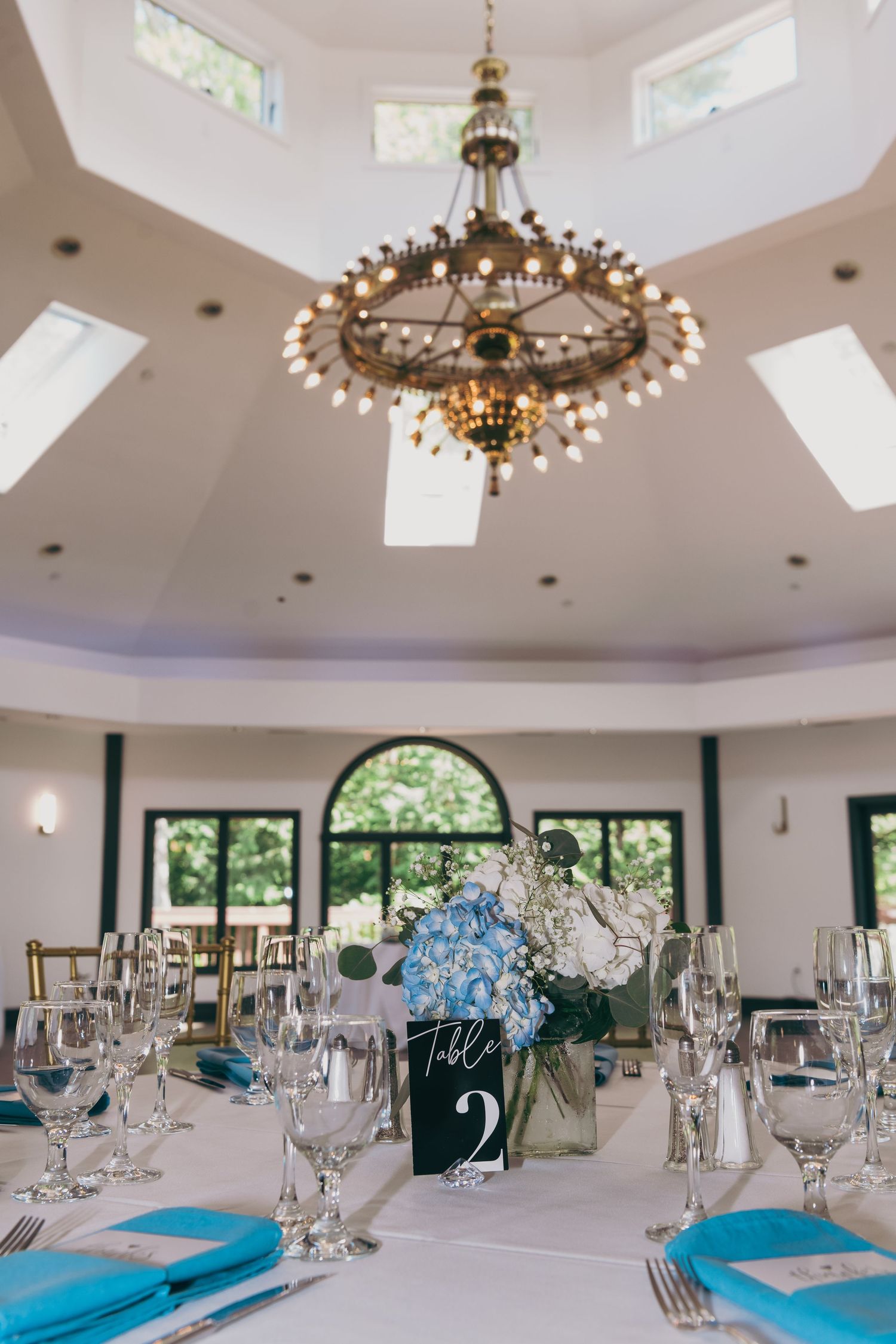 Elegant dining table setup with blue accents and floral centerpiece under a grand crystal chandelier in a bright circular room.