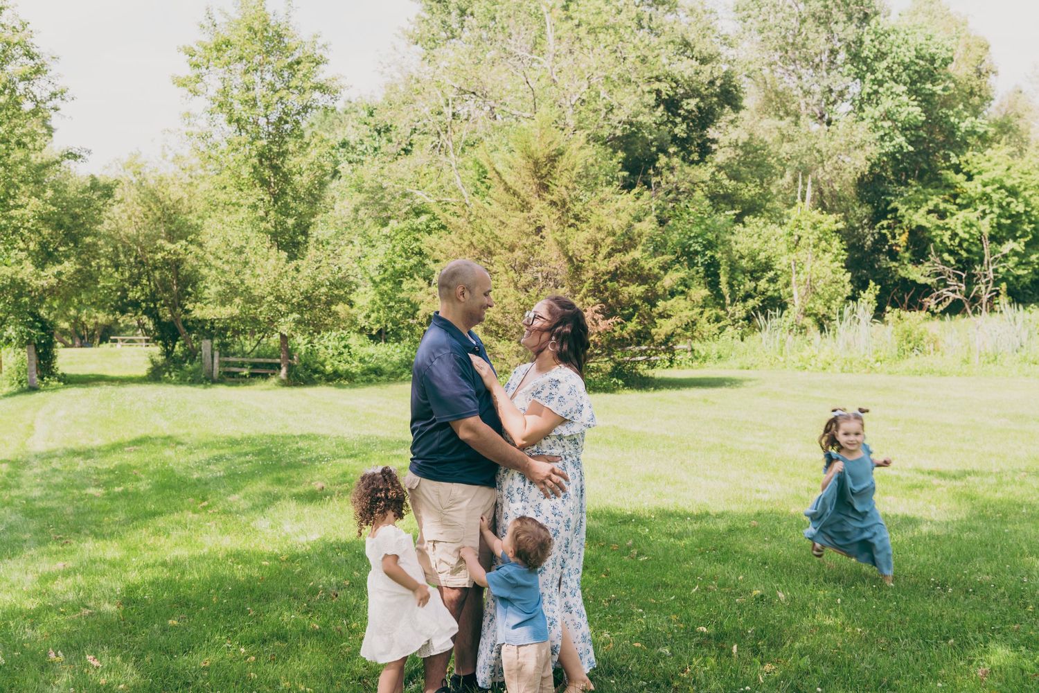 Children play together in a grassy field surrounded by trees on a sunny afternoon.