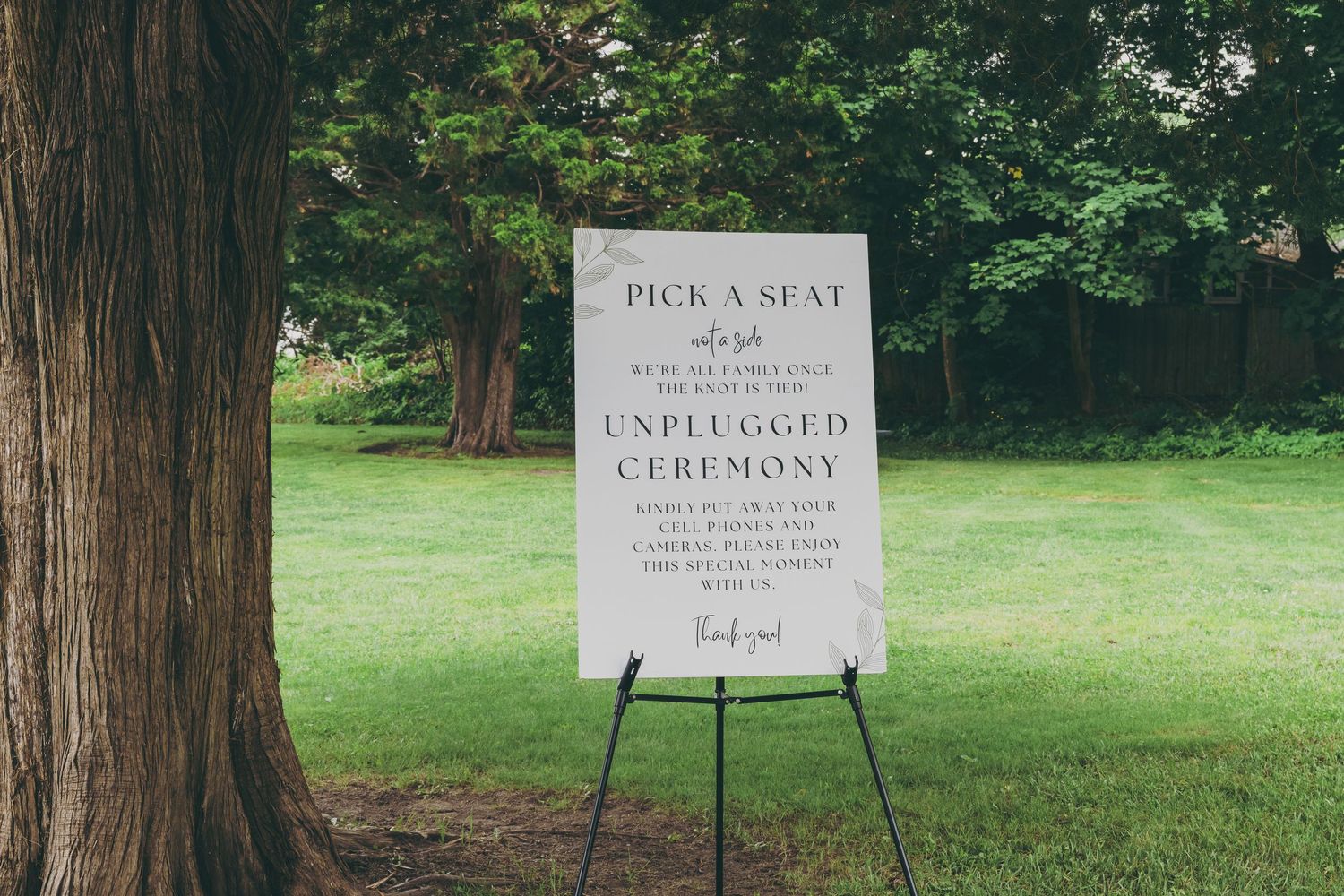 A rustic outdoor wedding venue with wooden welcome signs and lush greenery surrounded by tall trees.