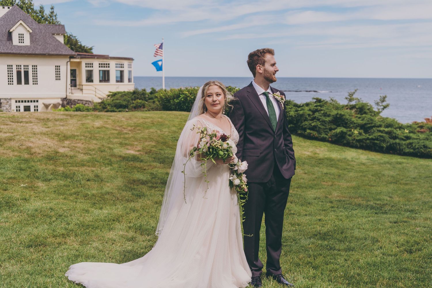 Couple in formal wedding attire stroll along coastal grass field on sunny day with ocean views.