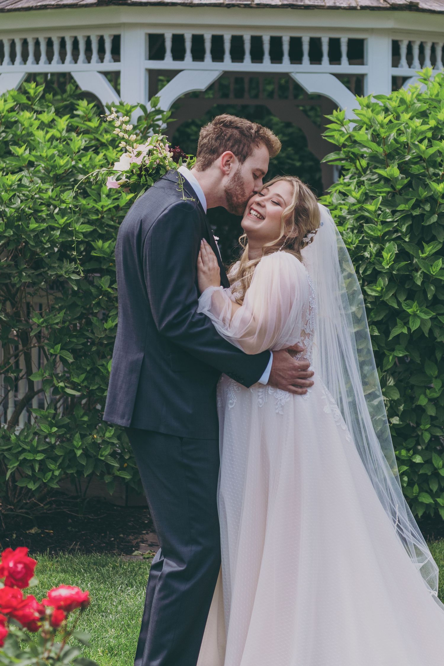 A tender wedding moment captured between newlyweds sharing kisses in a garden with greenery and red flowers.