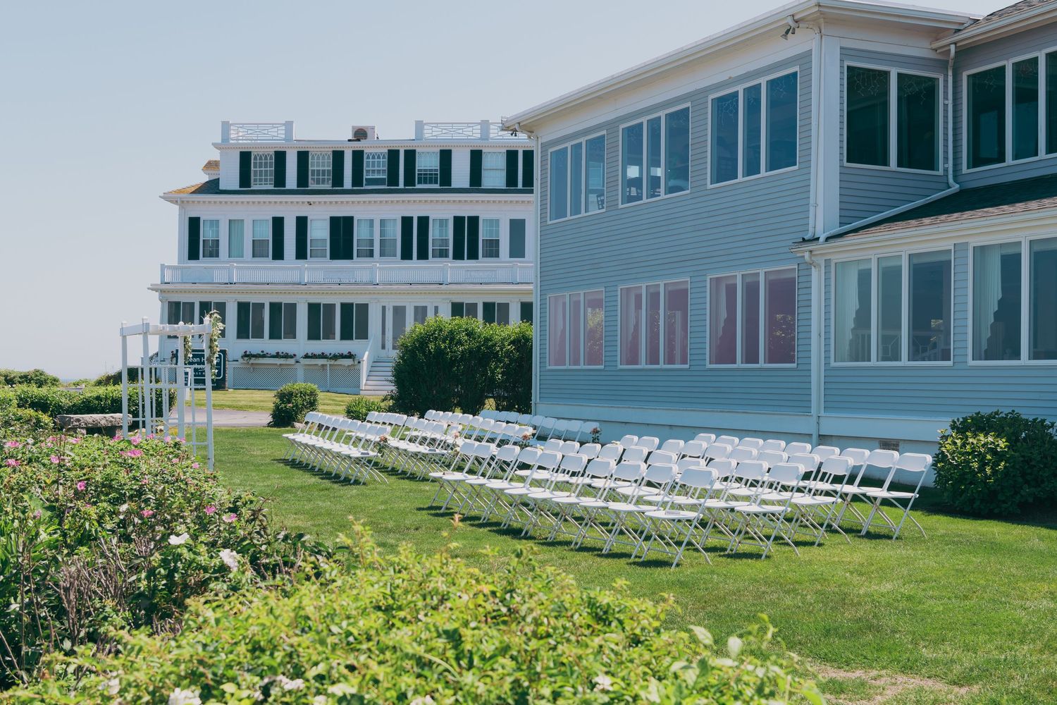 White folding chairs line the lawn outside a pale blue seaside hotel with large windows on a sunny summer day.