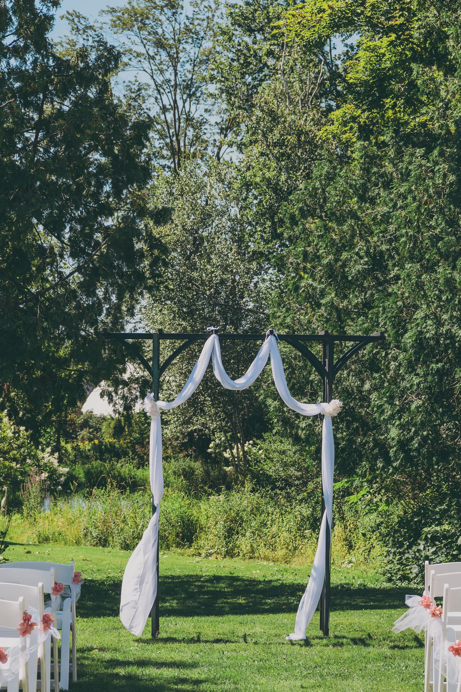 A wedding arch decorated with white fabric stands on a grassy lawn surrounded by trees in an outdoor ceremony setup.