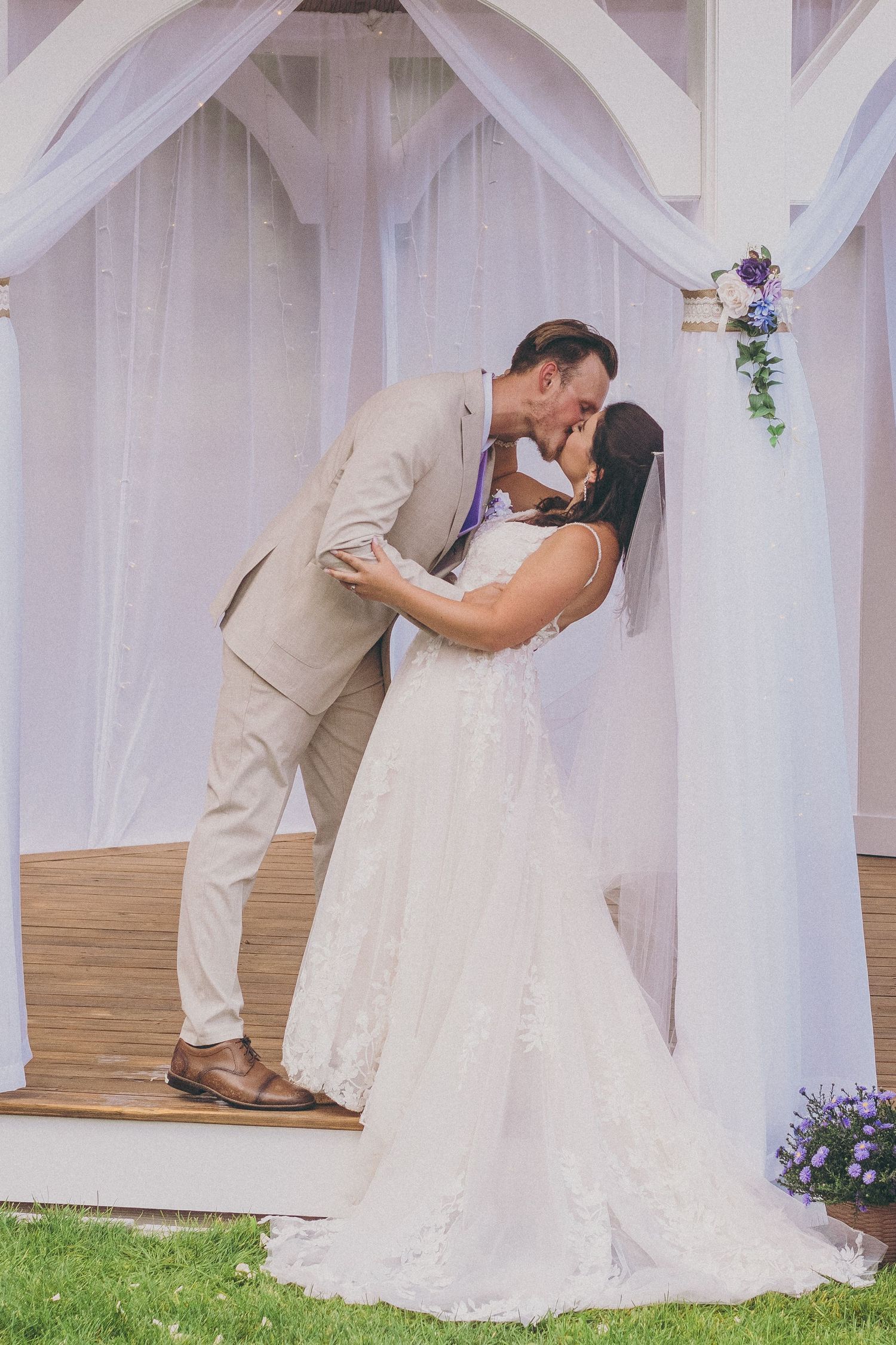 A couple shares their first kiss as newlyweds under a white fabric draped arch during an outdoor wedding ceremony.