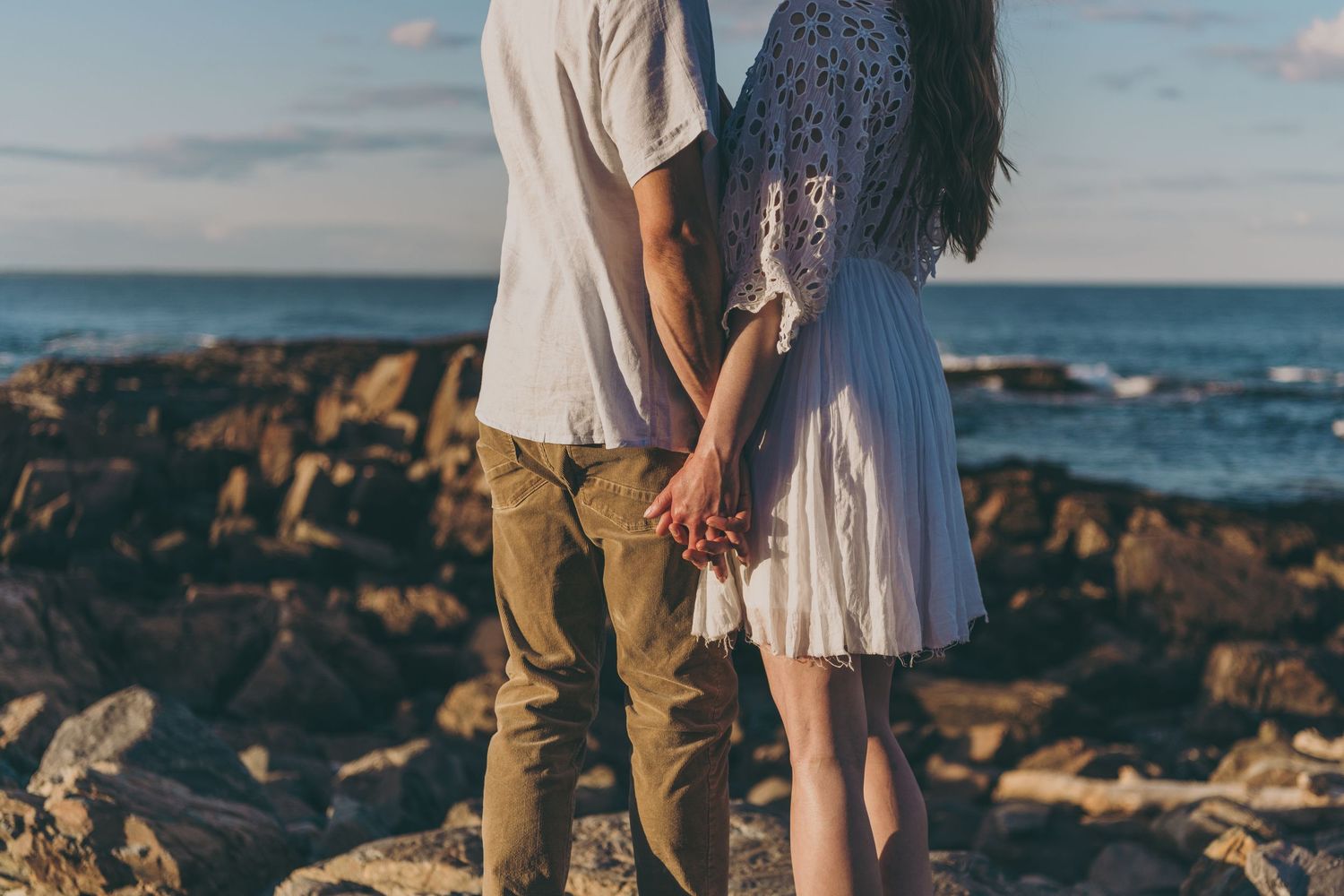 A couple holds hands while standing on rocky coastal shore at sunset with ocean waves in background.