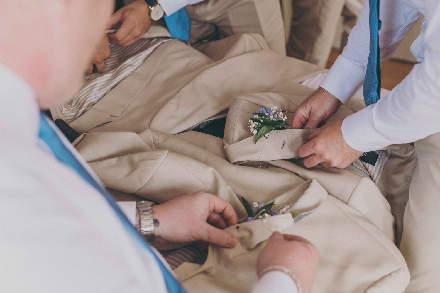 Hands pinning boutonniere flowers made of white blooms and greenery onto beige suit jacket.