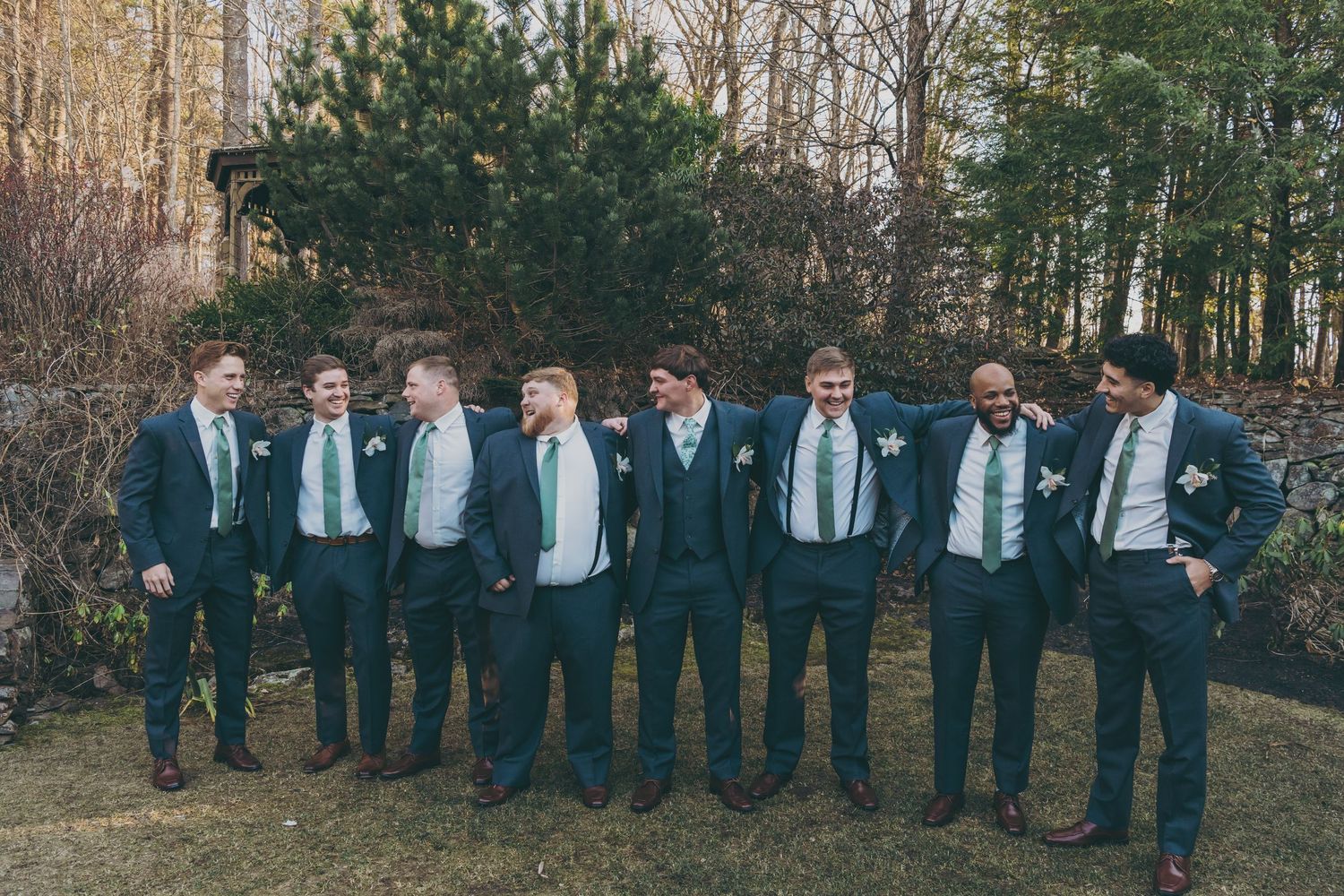 Groomsmen in matching navy suits and ties pose together outdoors in front of pine trees.