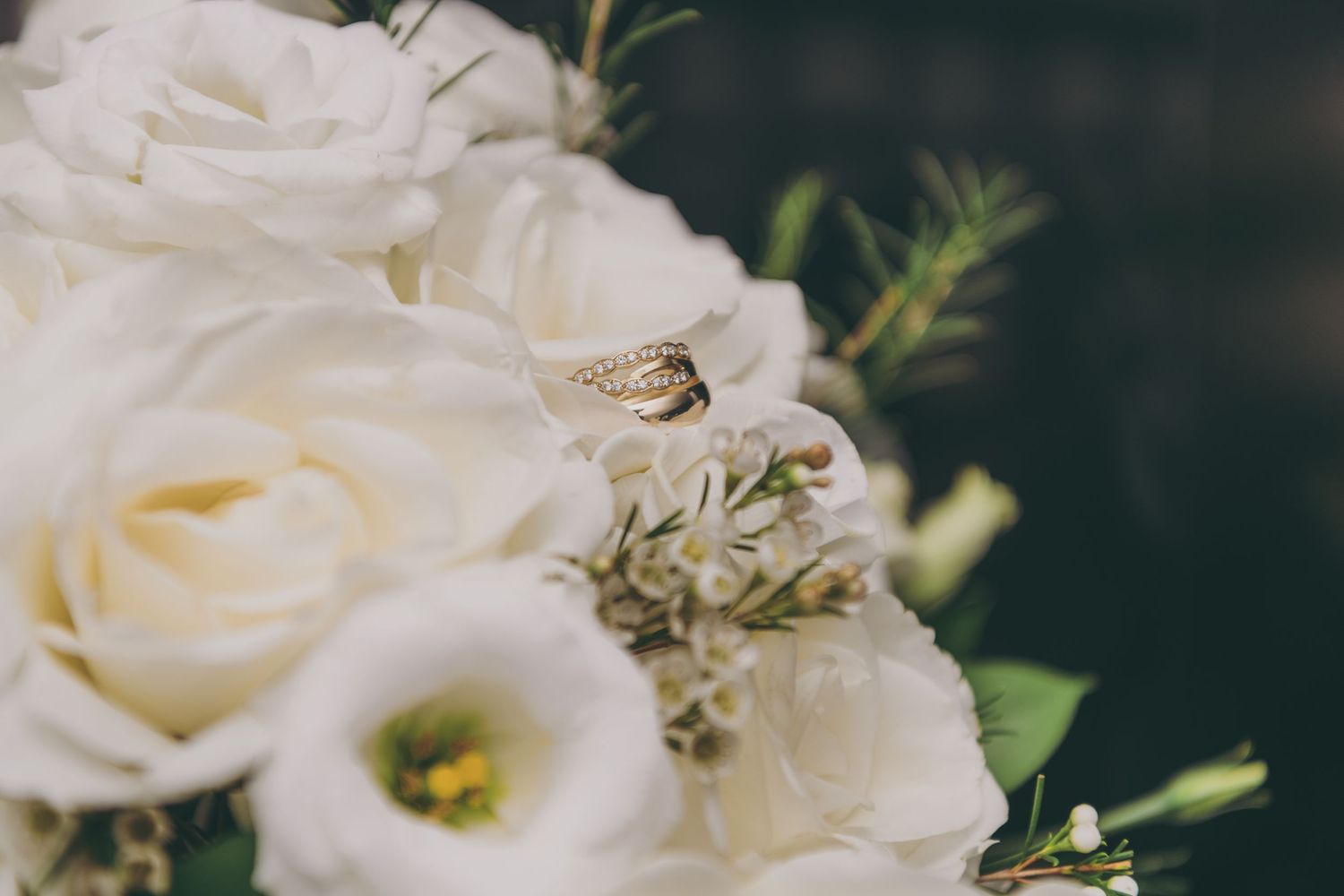 A delicate wedding ring rests atop elegant white roses and greenery in a romantic bridal bouquet.