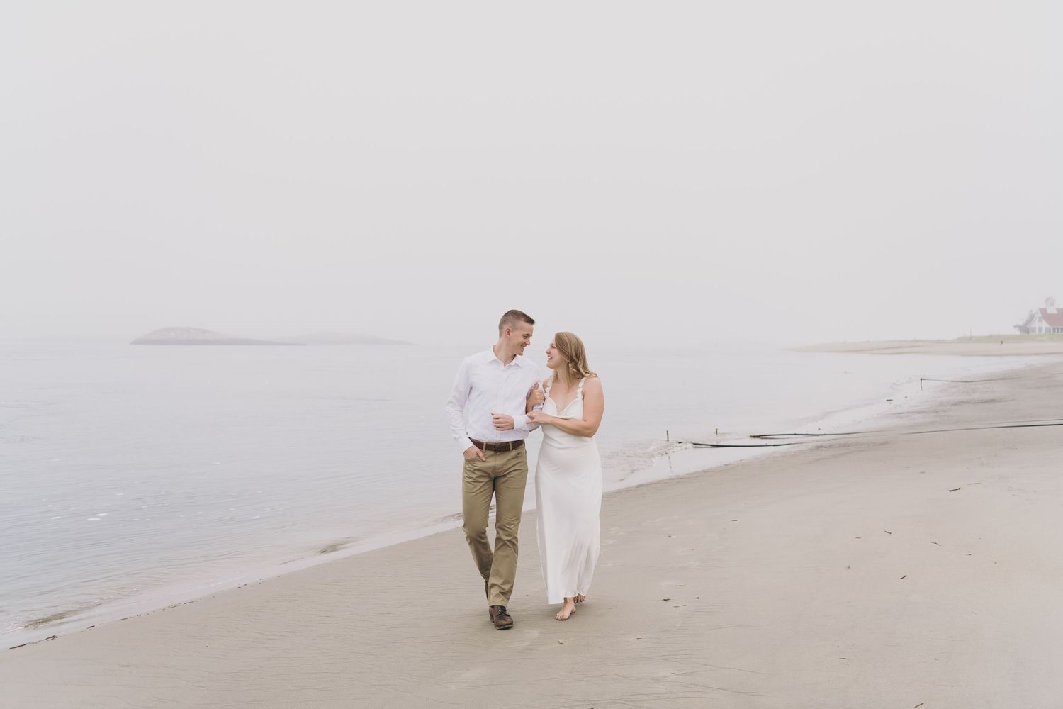A couple walks hand in hand along a misty beach shore wearing casual white and beige attire.