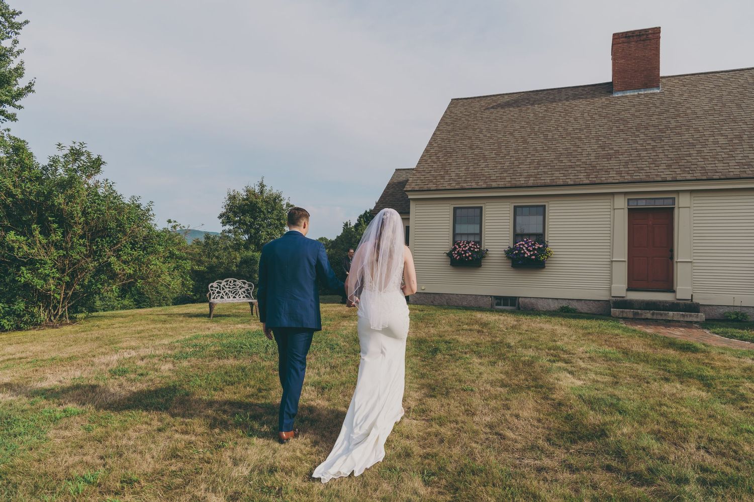Couple walks together on grass near colonial-style house during wedding day.