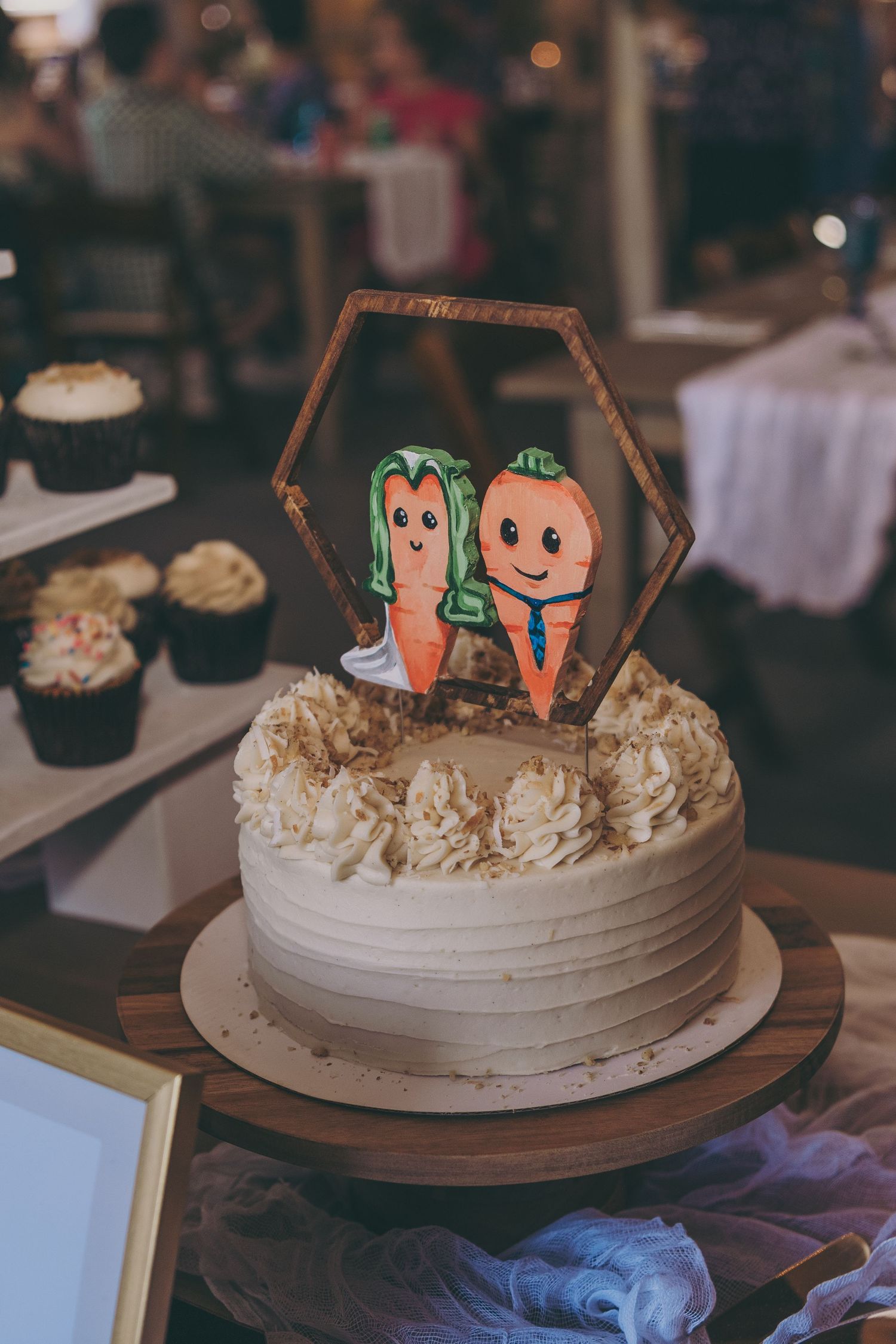 White frosted wedding cake topped with cute carrot couple cake topper in hexagon frame.