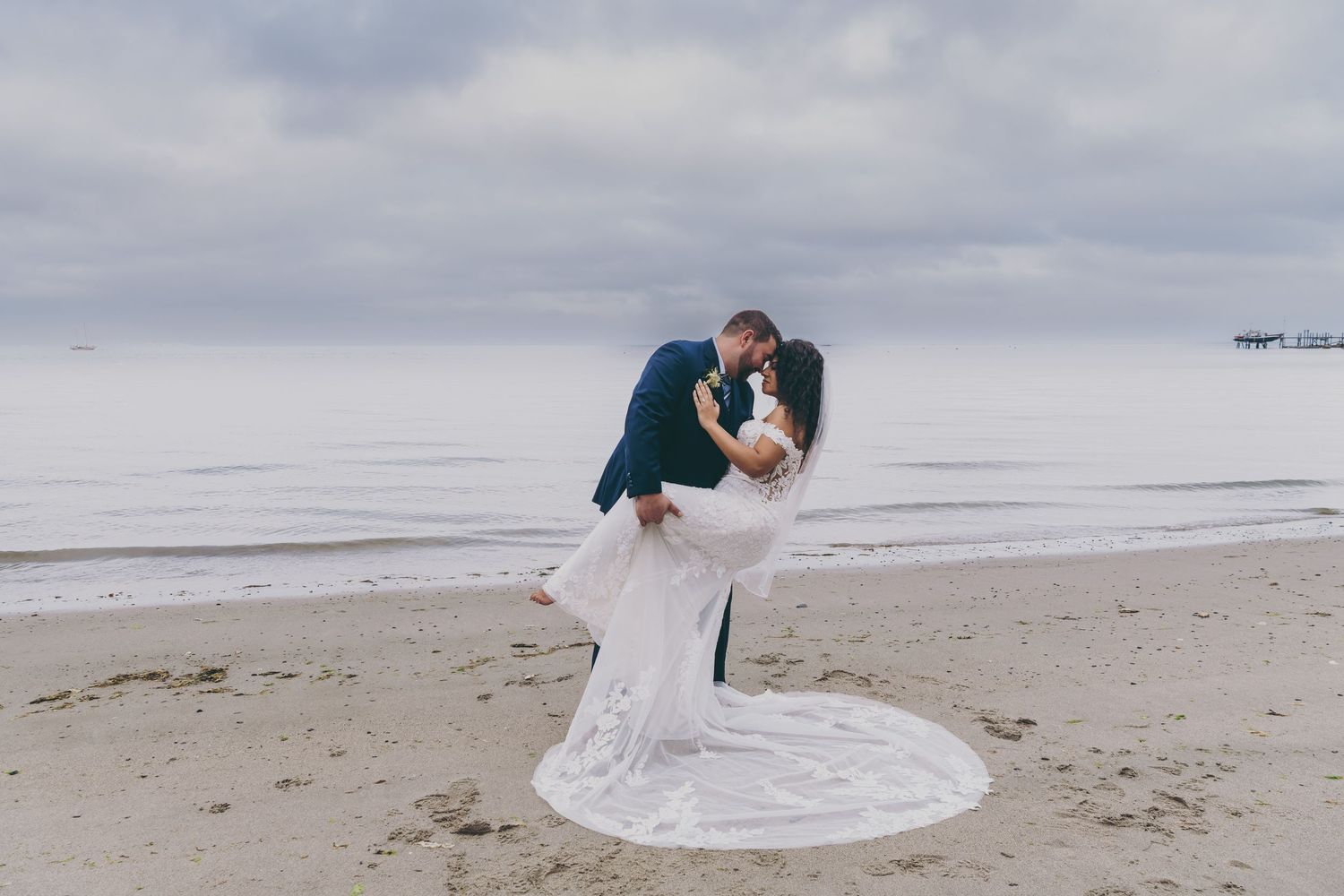 Elegant beach wedding couple dancing and embracing on a sandy shore under cloudy coastal skies.