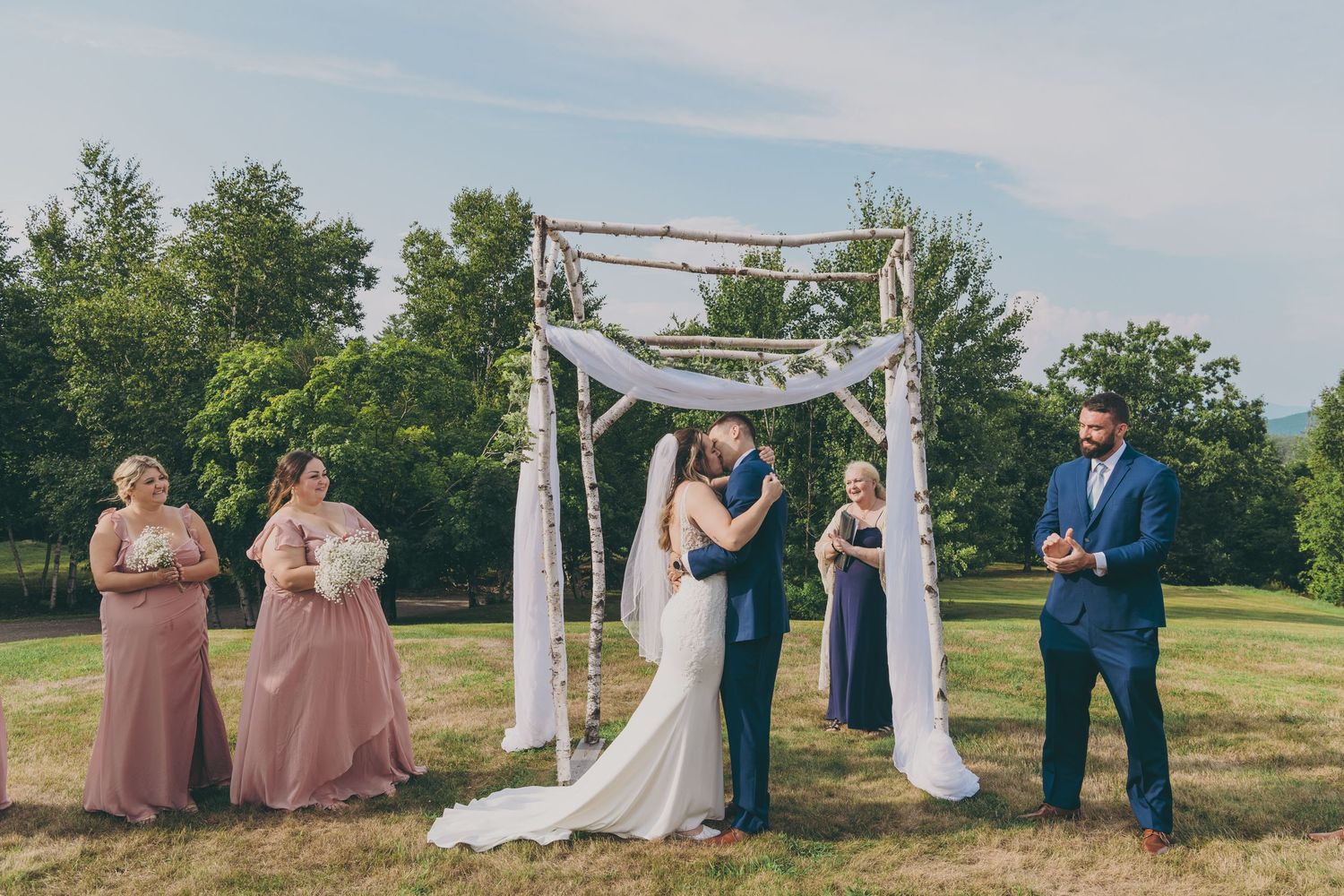 Outdoor wedding ceremony under a wooden arch decorated with white fabric and greenery on a sunny summer day.
