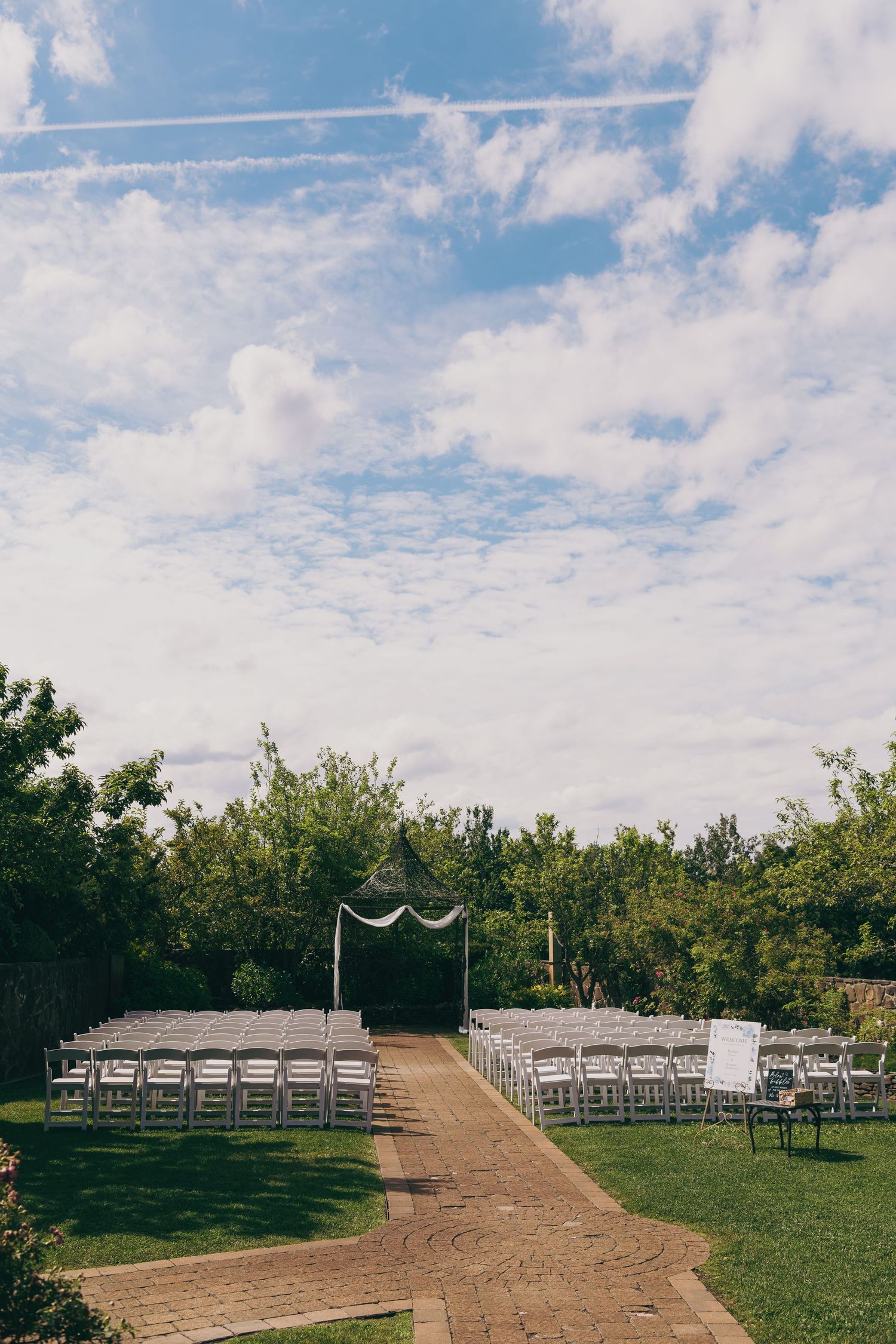 Outdoor wedding ceremony setup with white chairs and arch on dirt path surrounded by trees under blue cloudy sky.