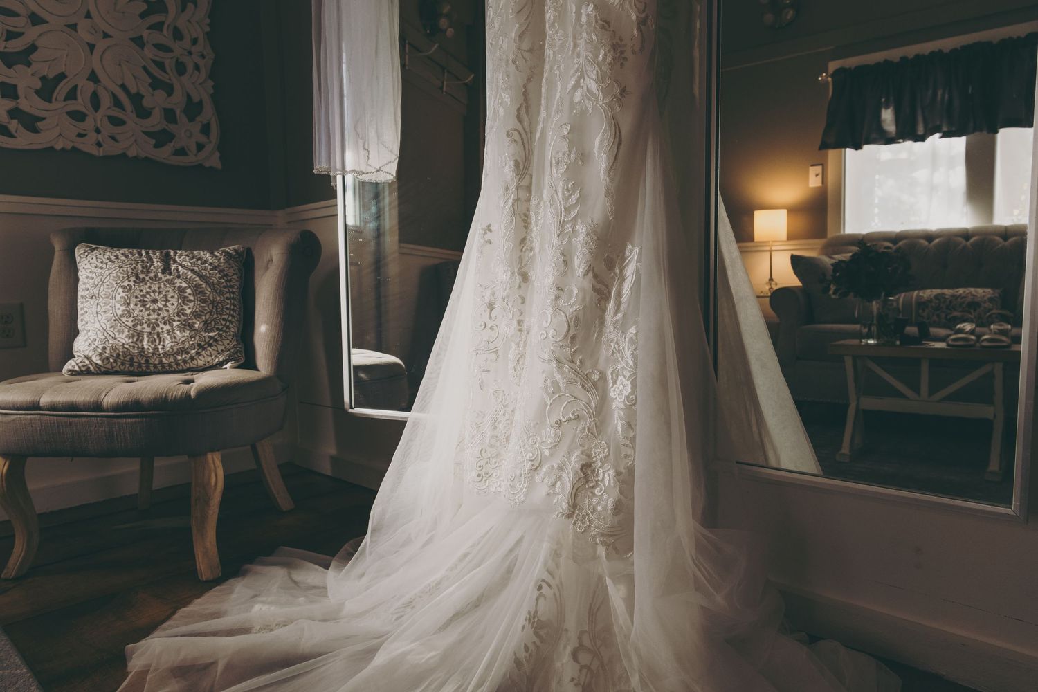 Elegant white lace wedding dress hanging in a room with vintage decor and soft lighting.