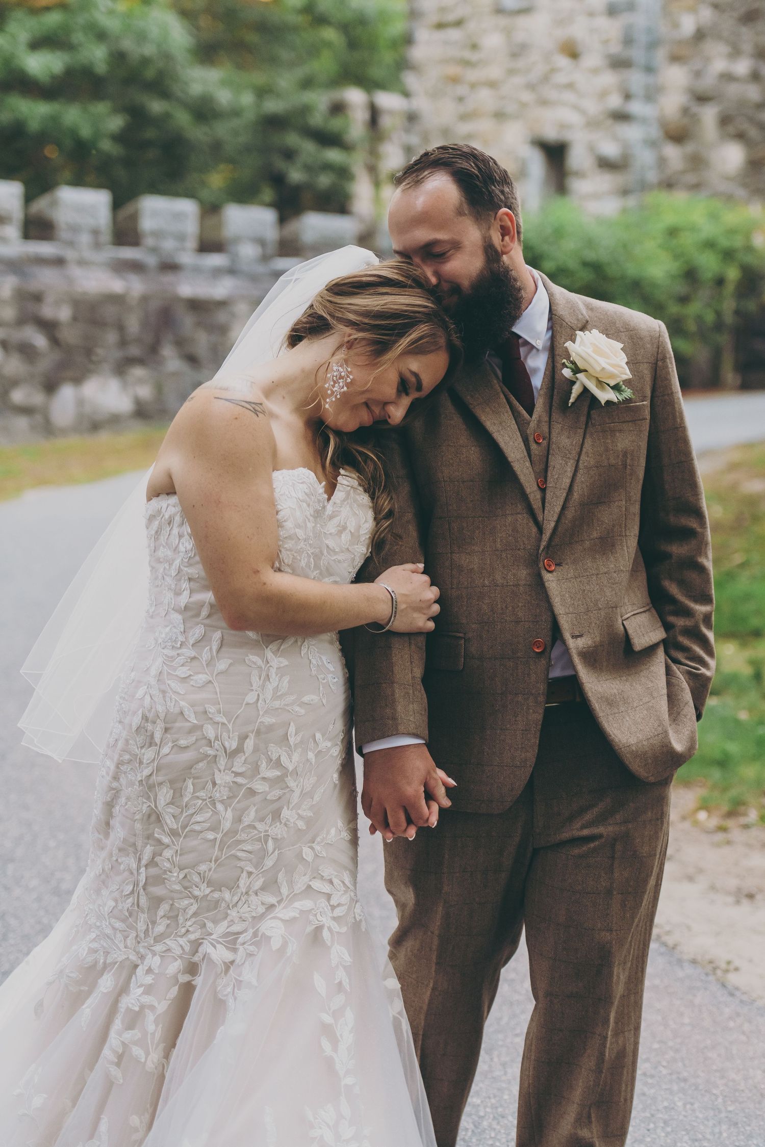 A tender wedding moment as newlyweds share an intimate embrace in front of a stone wall, bride in lace and groom in brown suit.