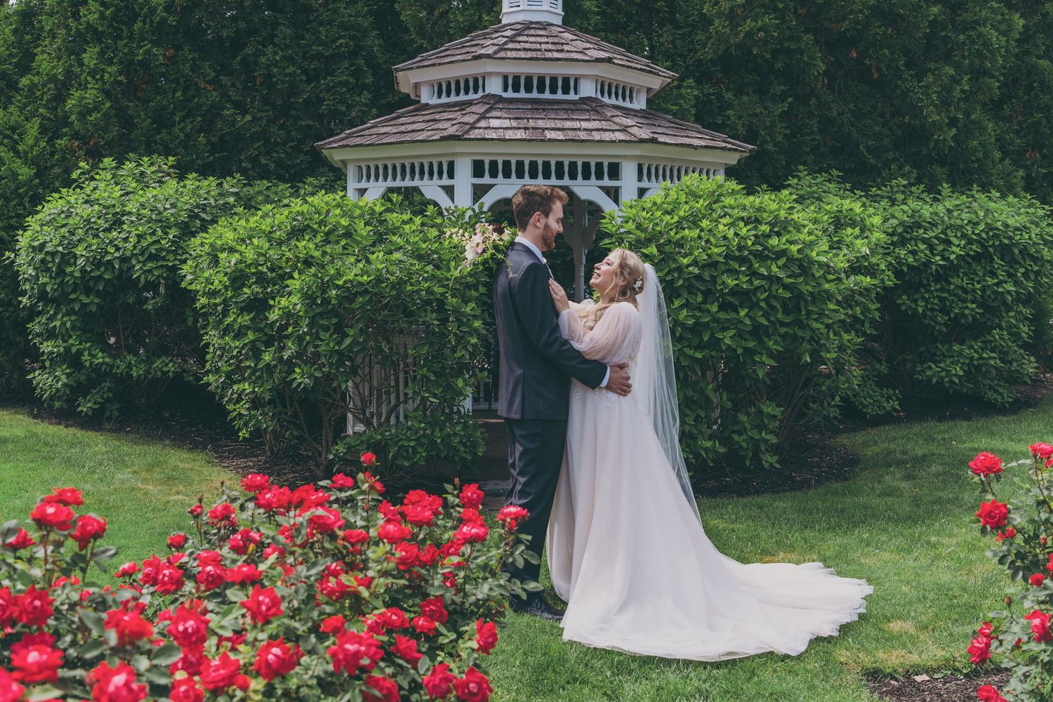 Wedding couple shares romantic moment in lush garden setting with white gazebo and blooming red roses.